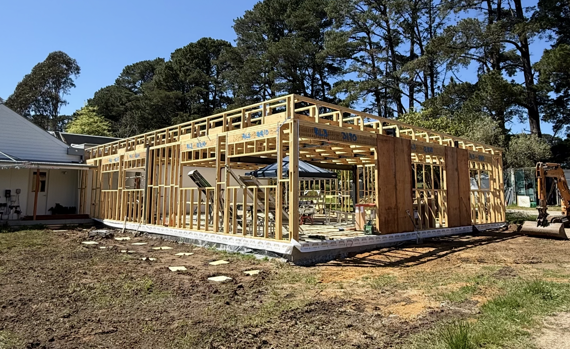 Building under construction with exposed wooden framing on a sunny day, with trees in the background.