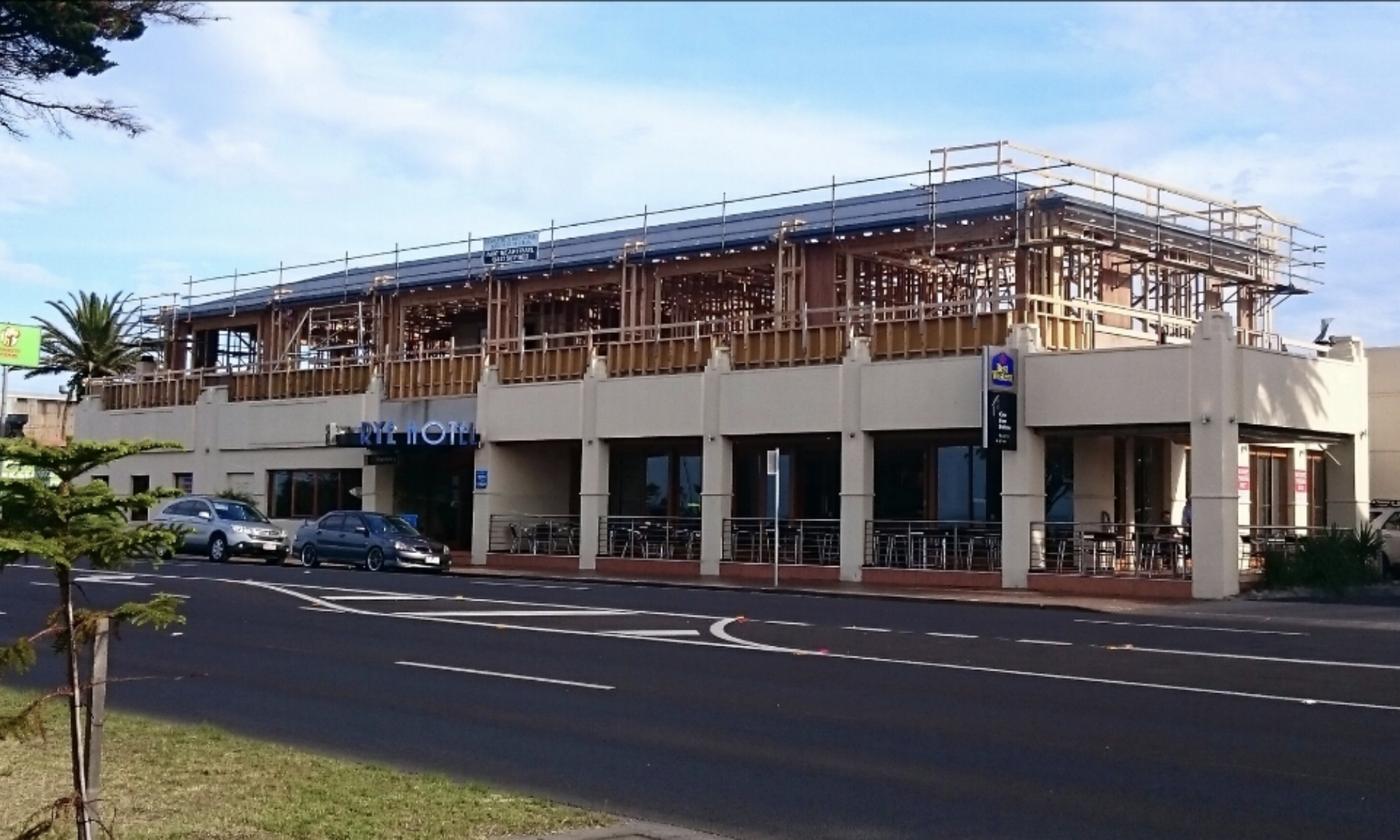 A two-story building under construction with a beige exterior and wood framework on the upper level, located in a commercial area with parked cars and a street in front.