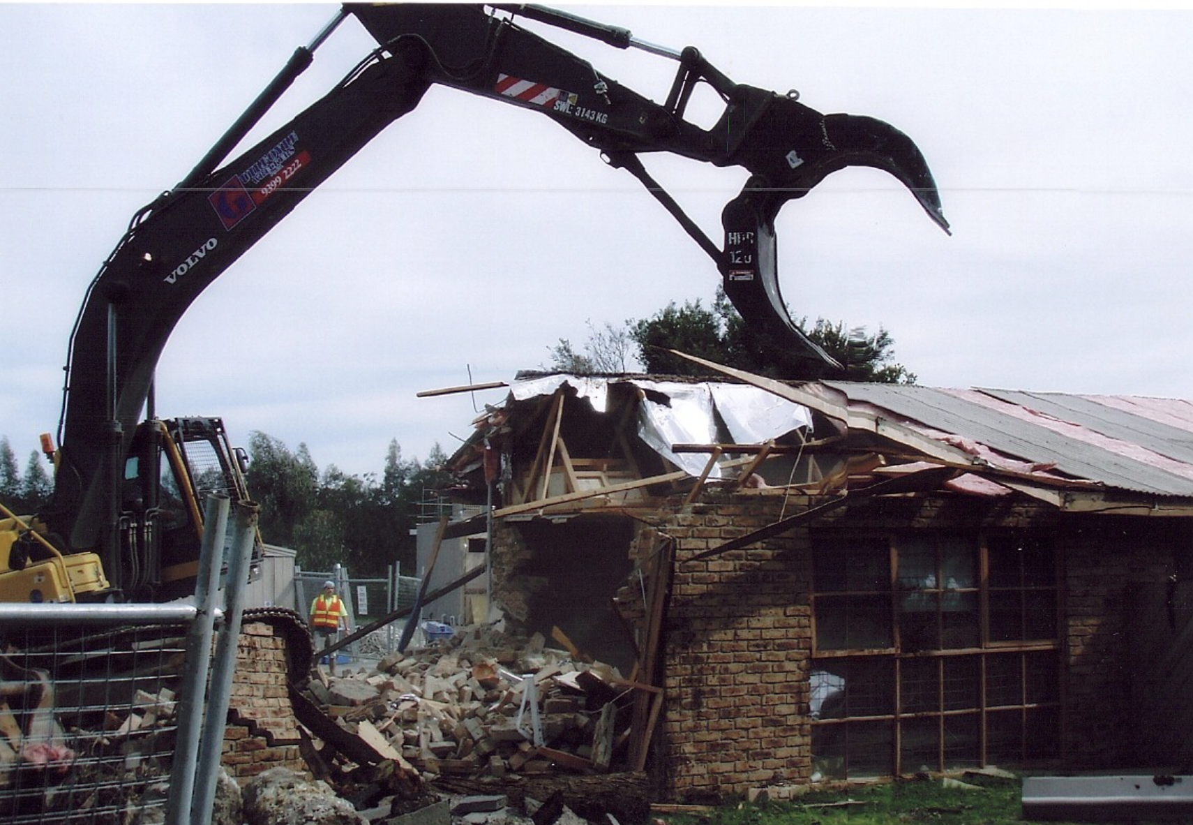 Construction site where a large excavator is demolishing a brick building, debris scattered, workers in safety vests and helmets present.