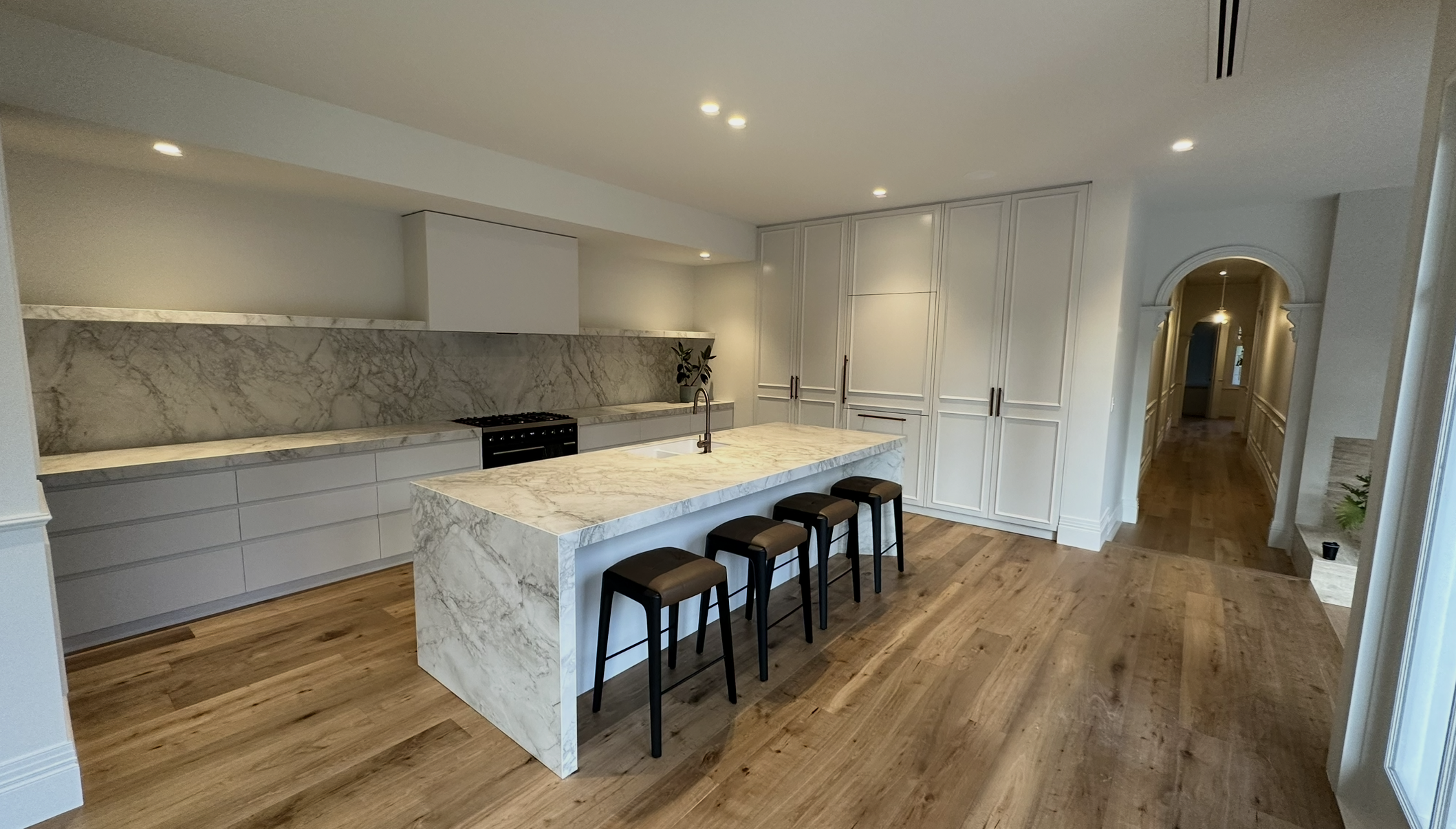 Modern kitchen with white cabinets, marble countertops, a marble kitchen island with four stools, wooden flooring, and a hallway in the background.