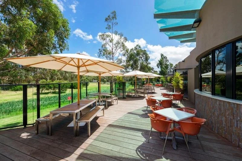 Outdoor patio with wooden tables, orange chairs, large white umbrellas, on a wooden deck, overlooking a green landscape with trees, under a blue sky with some clouds.