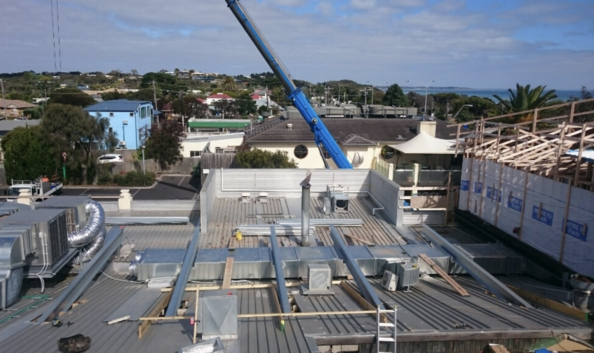 Construction site on a rooftops with a large blue crane and ongoing building framework, with suburban houses and ocean in the background.