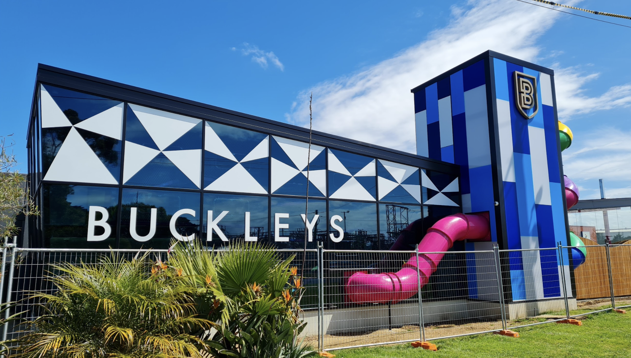 Exterior of a Buckley's store with blue geometric patterned facade, large glass windows, and a colorful water slide. The store has cool tones, a tall blue and white striped tower with a 'LB' logo, and is surrounded by a metal fence and landscaping.