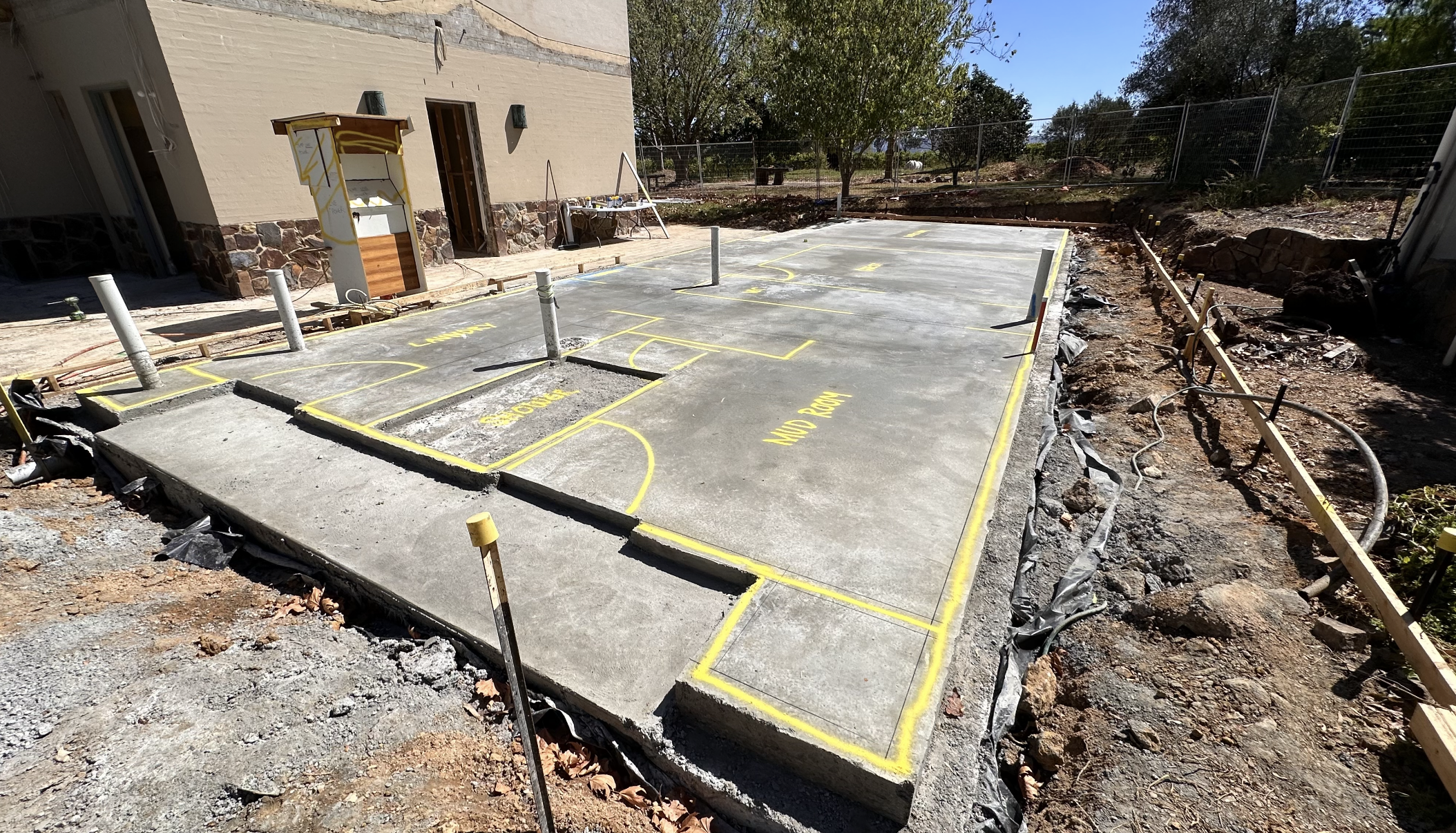 Concrete patio with basketball court markings, construction materials and tools nearby, surrounding dirt and trees in the background.