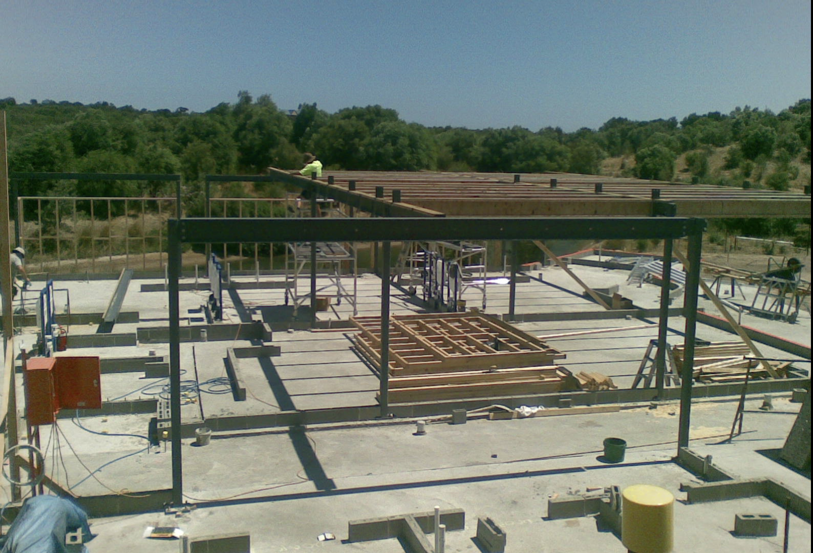 Construction site for a building with steel beams and wooden framing, workers on-site, surrounded by trees and a blue sky.