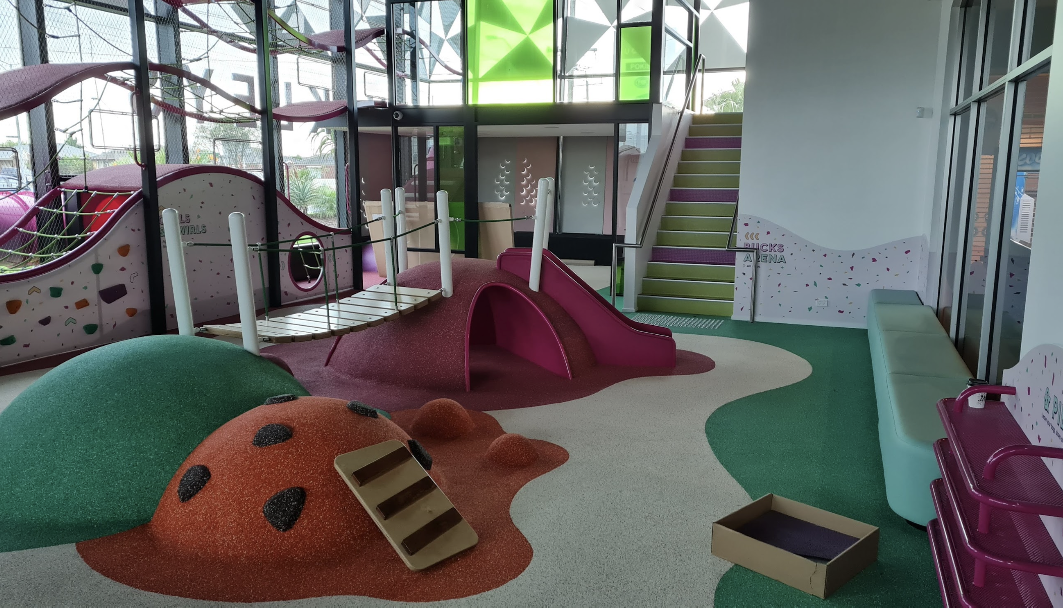 An indoor children's play area with colorful equipment including a pink slide, climbing structures, cushioned flooring, and seating.