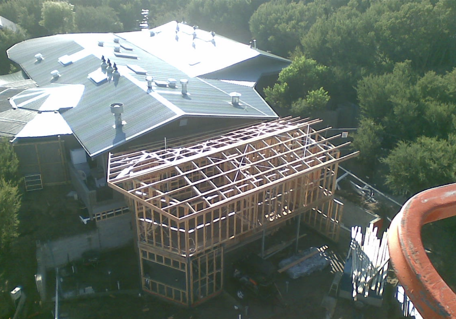 Aerial view of a construction site with a partially built wooden framework attached to an existing building with a metal roof surrounded by trees.