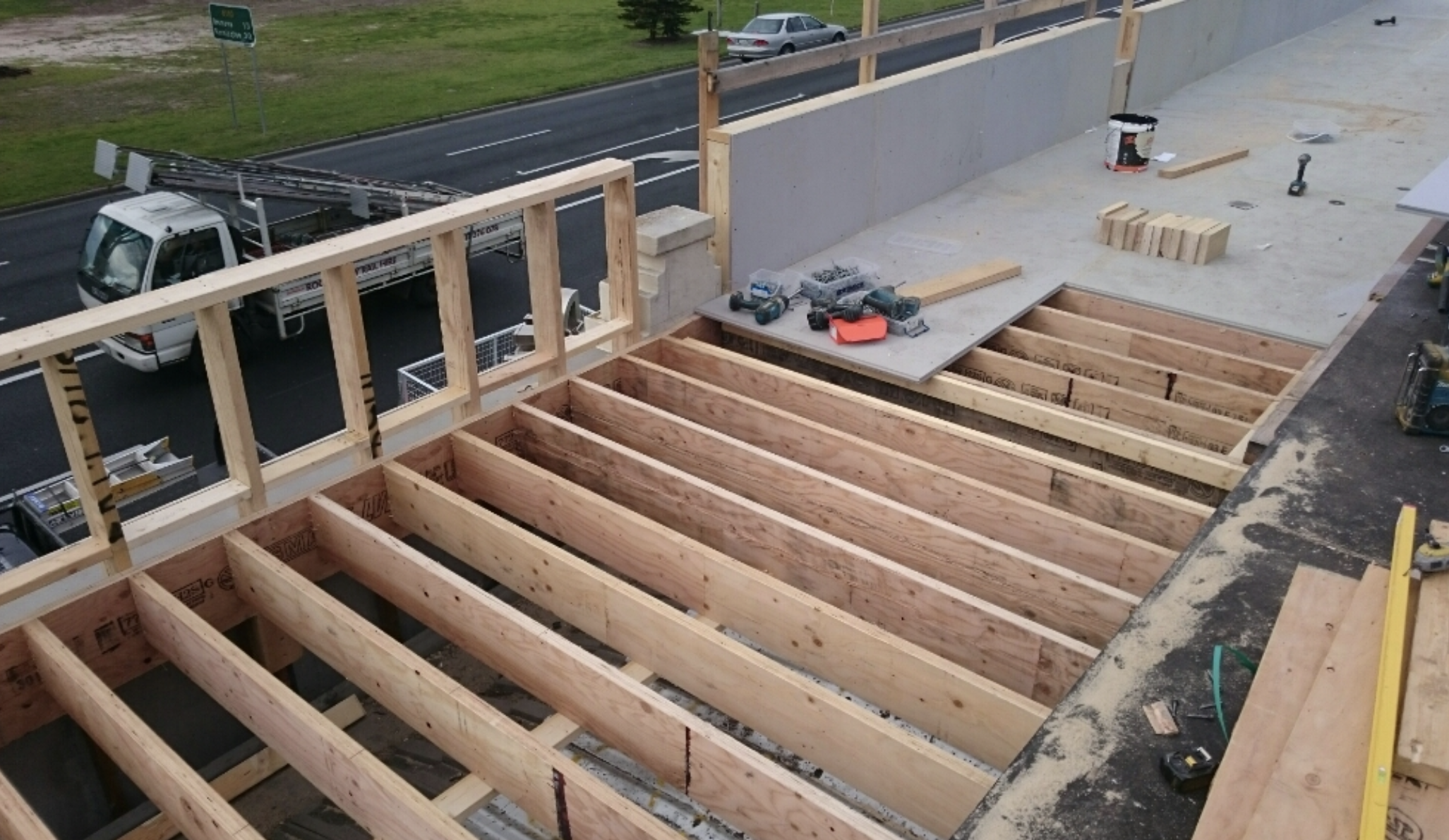 Construction site with wooden framing on upper floor, tools, and materials, overlooking a street with a truck and a grassy area.