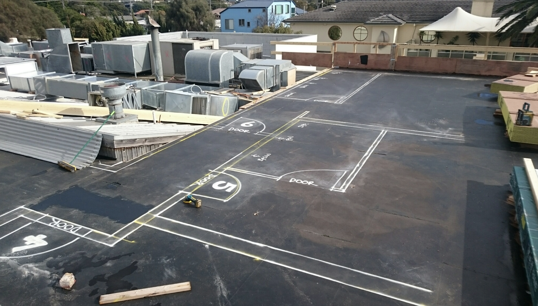 Roof construction site with white chalk markings indicating door placements and measurements, construction tools, stacks of insulation, and ventilation ducts, with residential buildings in the background.
