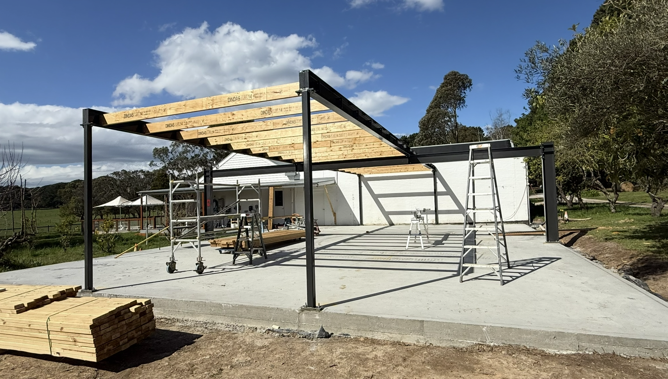 Construction site with a metal frame structure being assembled over a concrete slab, ladders, scaffolding, and building materials visible, in a rural area with trees and a house in the background under a partly cloudy sky.