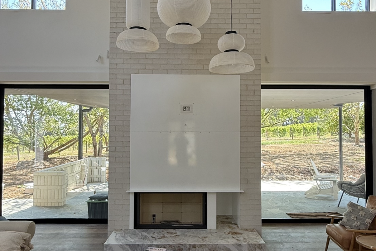 Interior view of a modern living room with a white brick fireplace, surrounded by large glass sliding doors that reveal a backyard with outdoor furniture and trees.