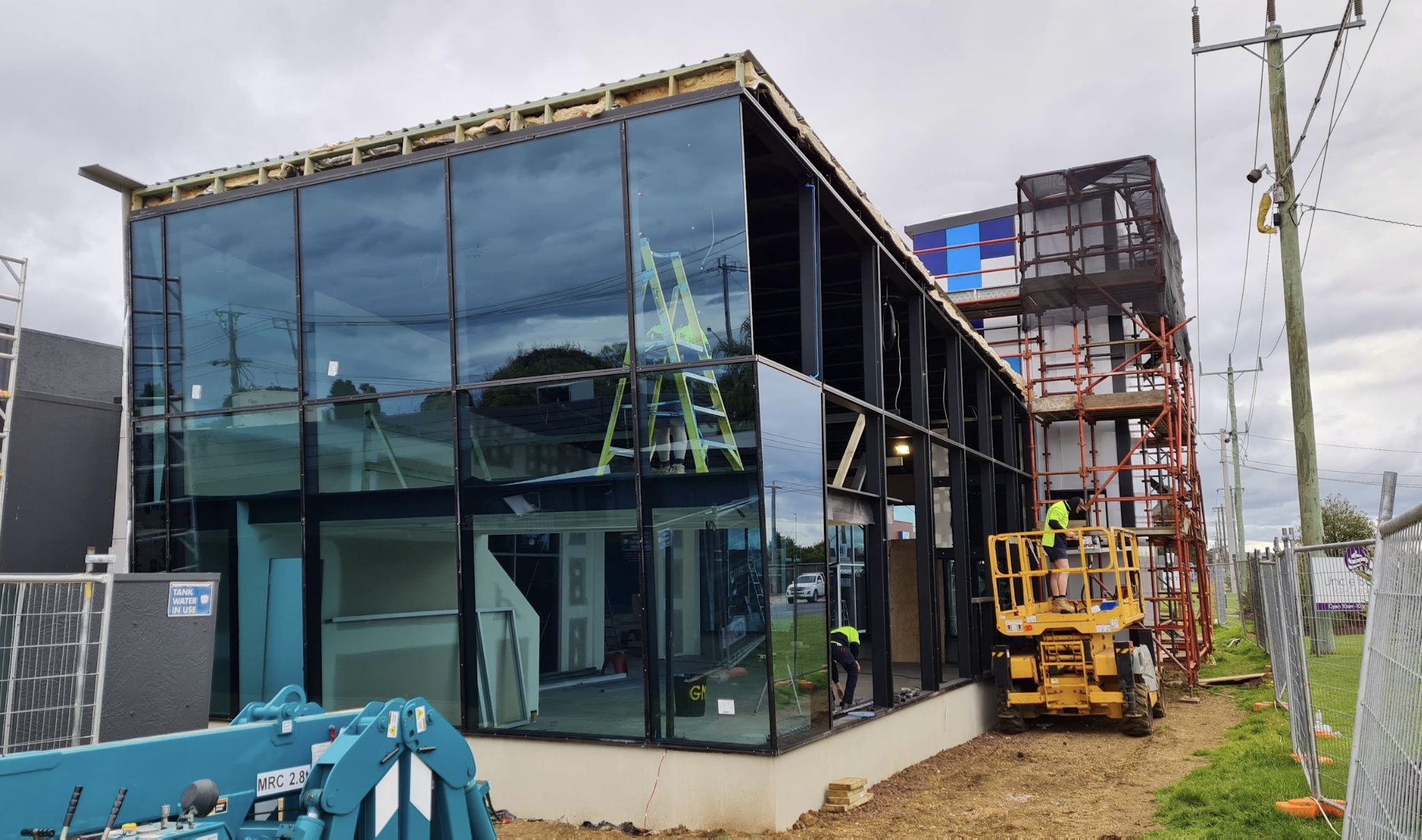 Construction workers building a modern glass-fronted commercial building, with scaffolding on the right side and a lift, during daytime with cloudy sky.