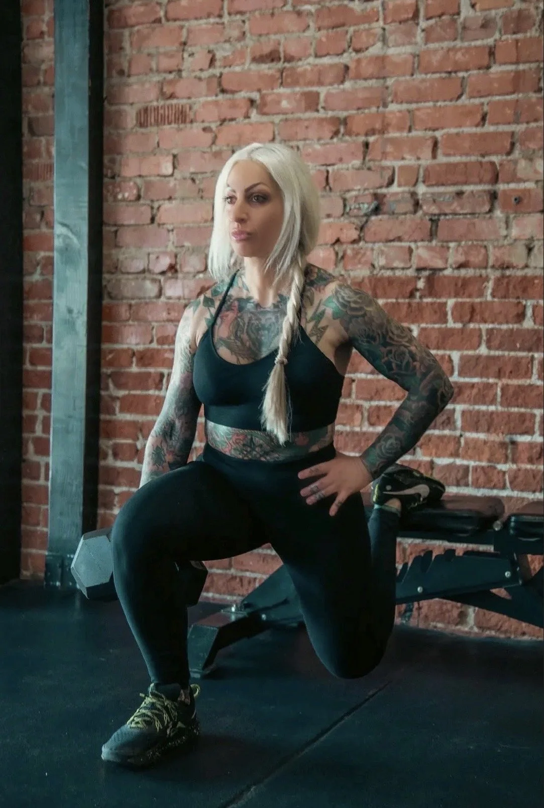 A woman with platinum blonde hair in a braid, tattoos on her arms and chest, dressed in black workout clothing, doing a lunge with one foot on a bench in a gym with a brick wall background.