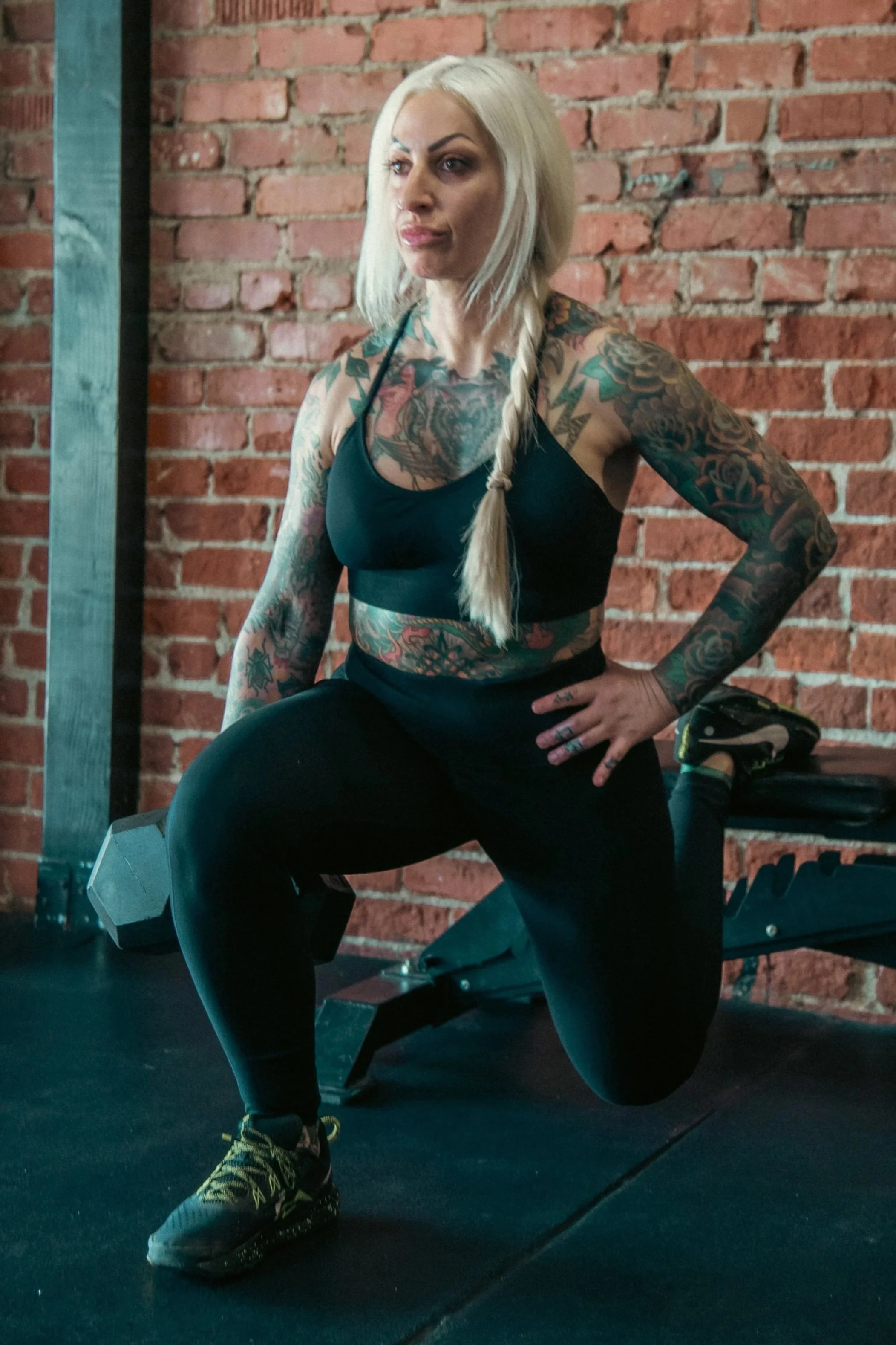 A woman with platinum blonde hair, tattoos on her arms and chest, wearing a black athletic top, black pants, and sneakers, is working out in a gym with a brick wall background. She is performing a squat with a dumbbell resting on her right thigh.