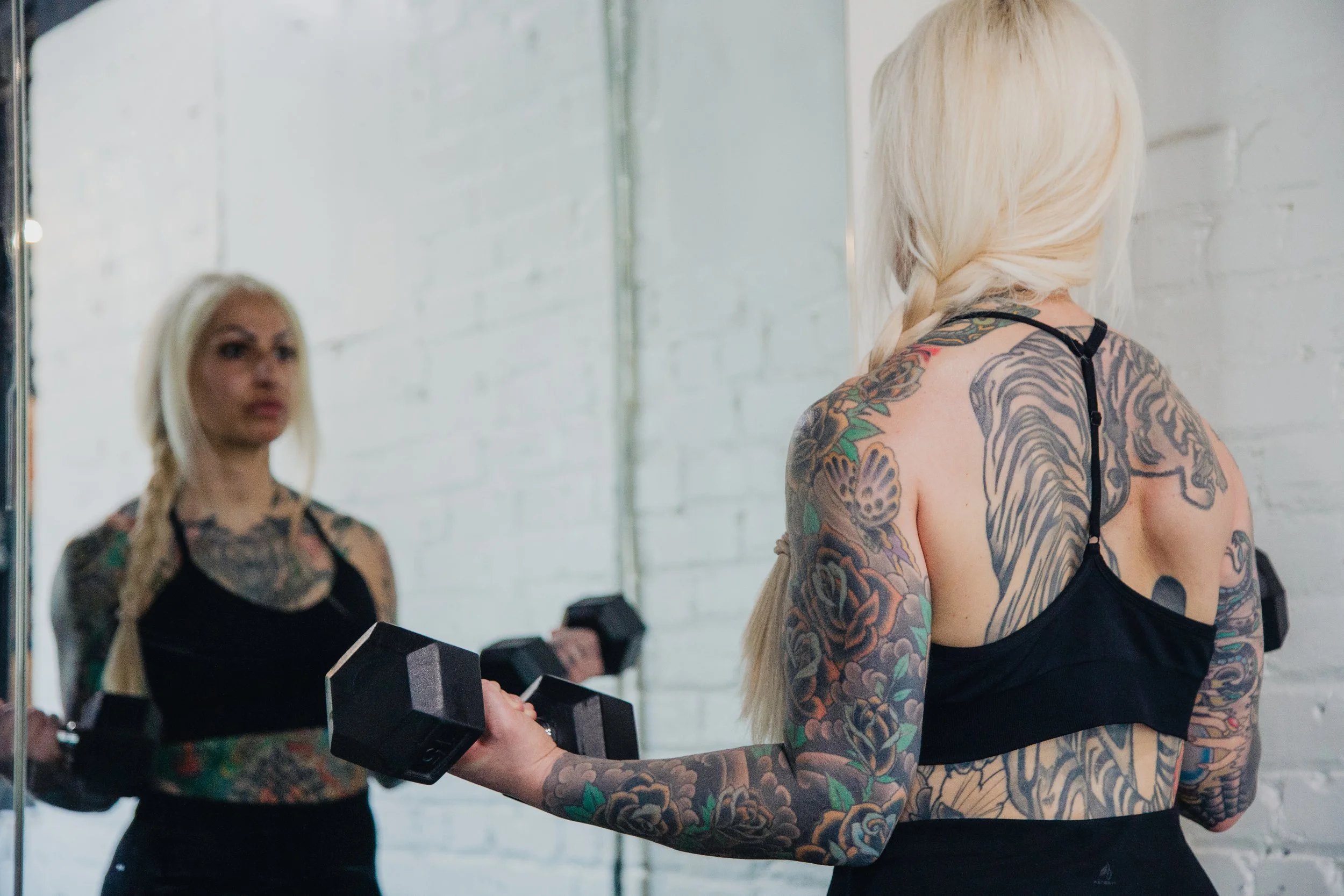 A woman with tattoos on her arms and back lifting dumbbells in a gym, with her reflection visible in a mirror.