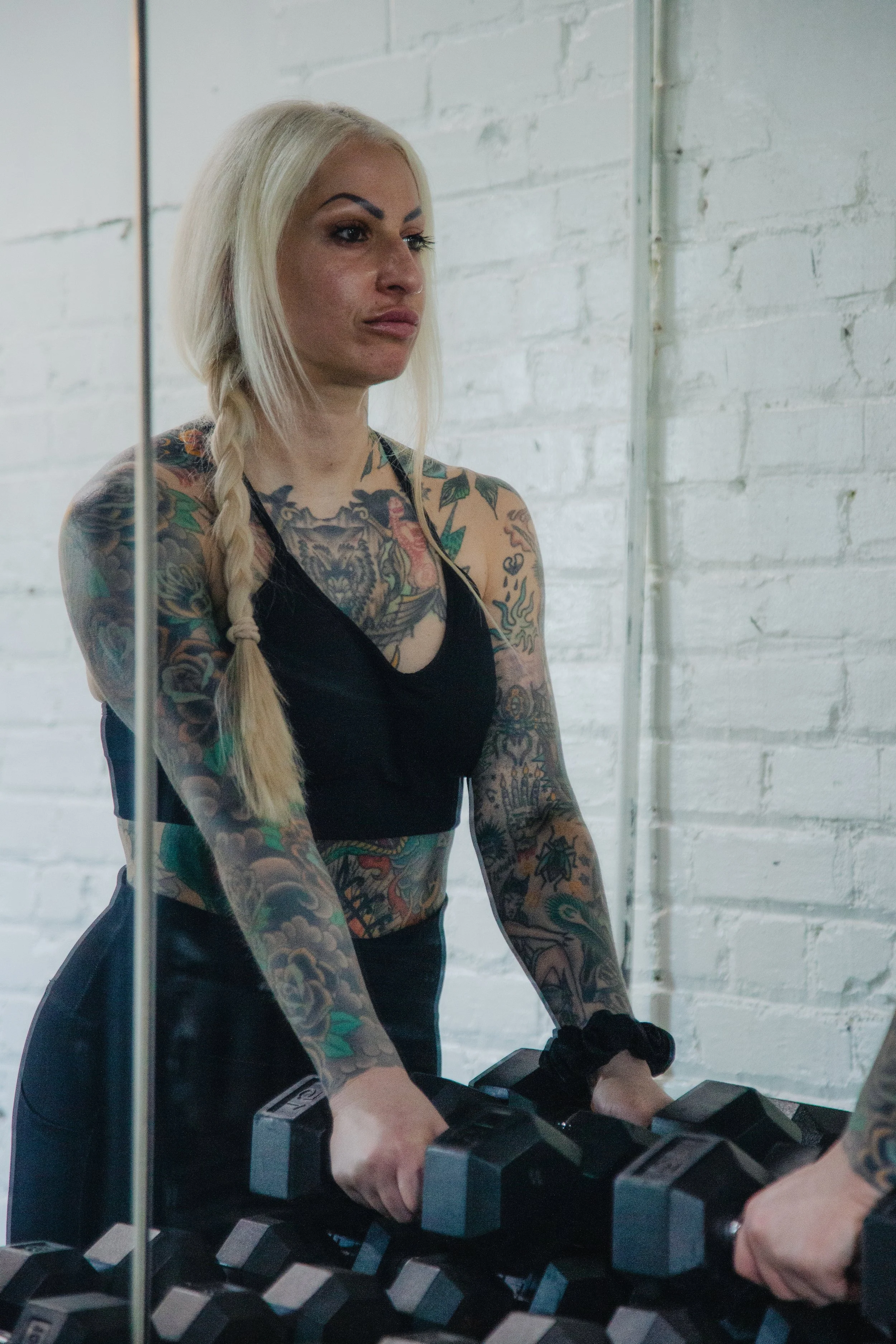 A woman with platinum blonde hair, tattoos covering her arms and chest, wearing a black tank top, standing in a gym and lifting dumbbells.