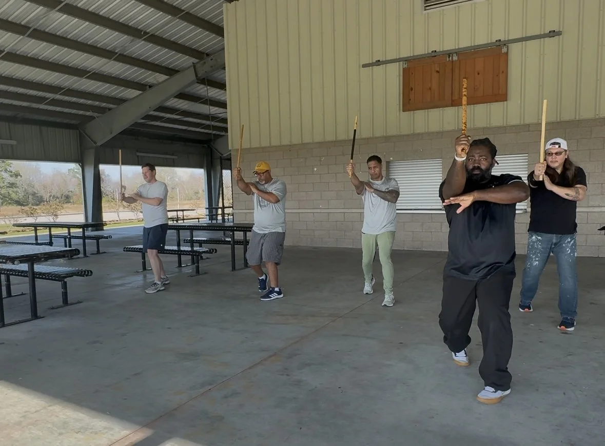 Five men practicing martial arts moves with sticks inside a covered outdoor pavilion.