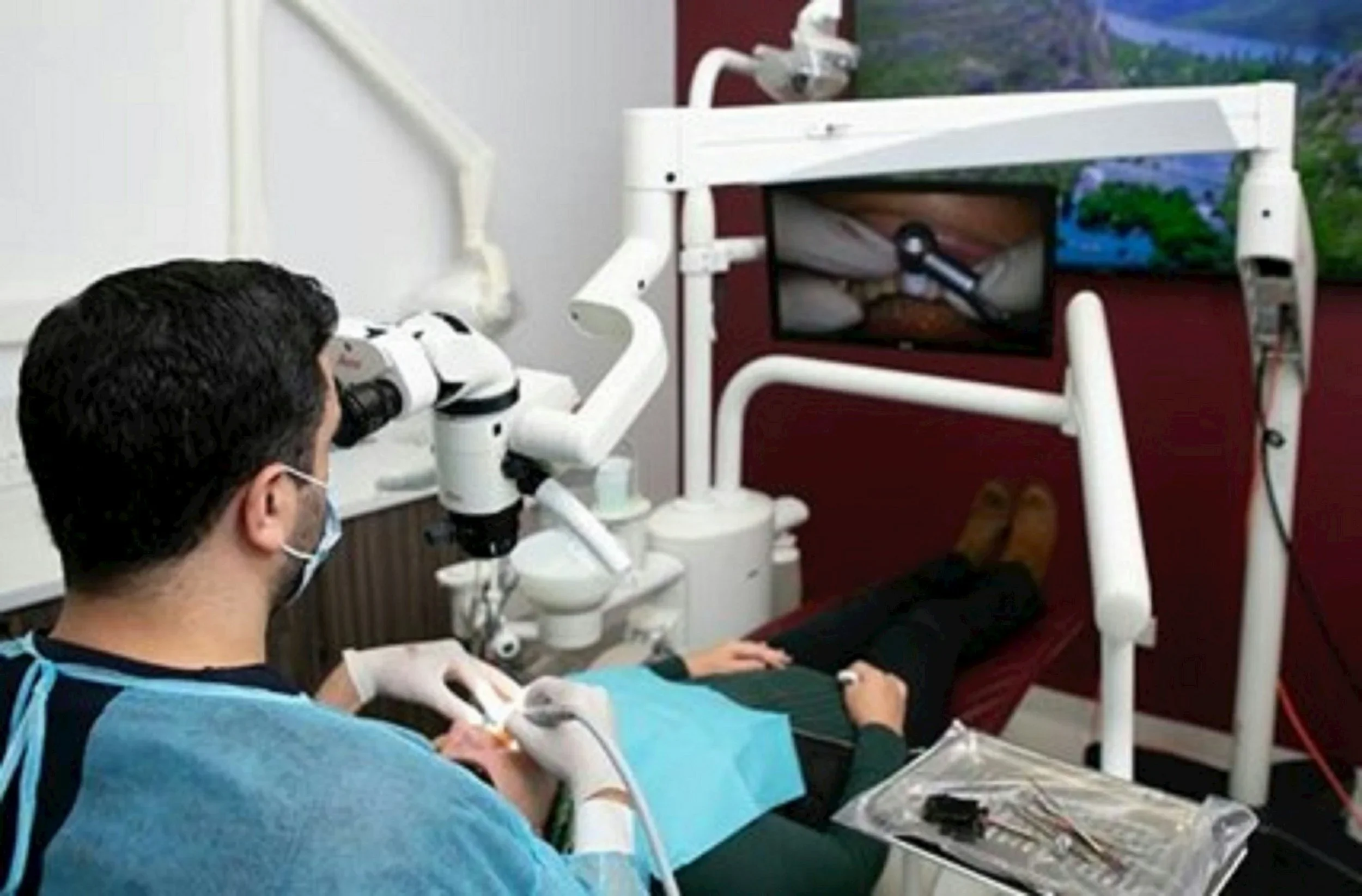 A medical professional in scrubs, mask, and gloves using a microscope for a procedure on a patient lying on a specialized dental or surgical chair in a clinic. The procedure involves a small tool or instrument, and there is a tray with medical instruments nearby.