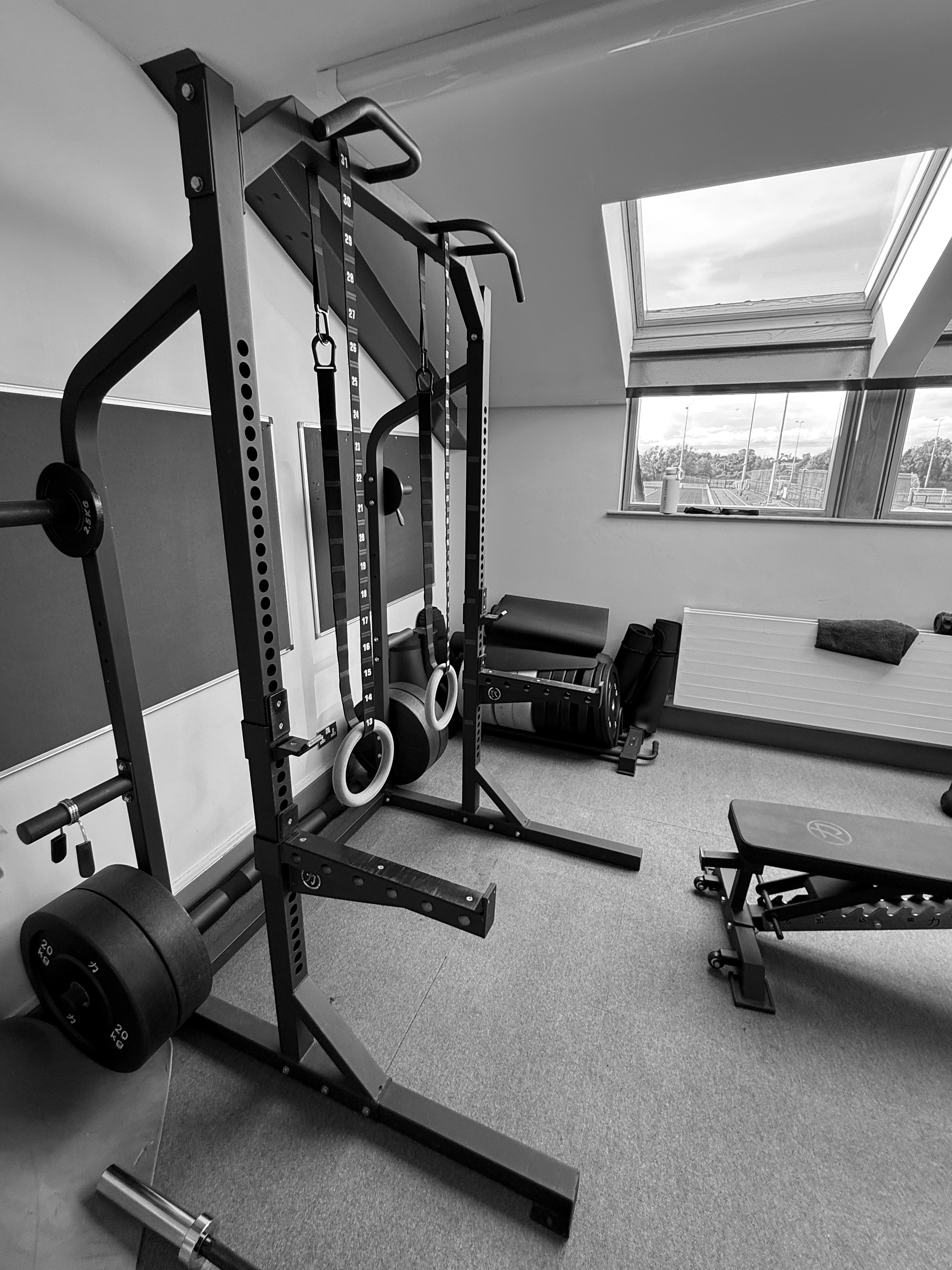 Home gym with squat rack, gymnastic rings, exercise mat, roller, and ventilation at the window.