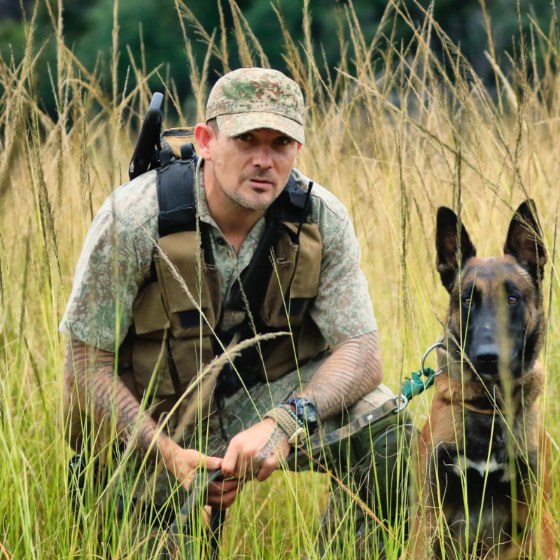 Man in camouflage outdoors with a German Shepherd dog in tall grass.