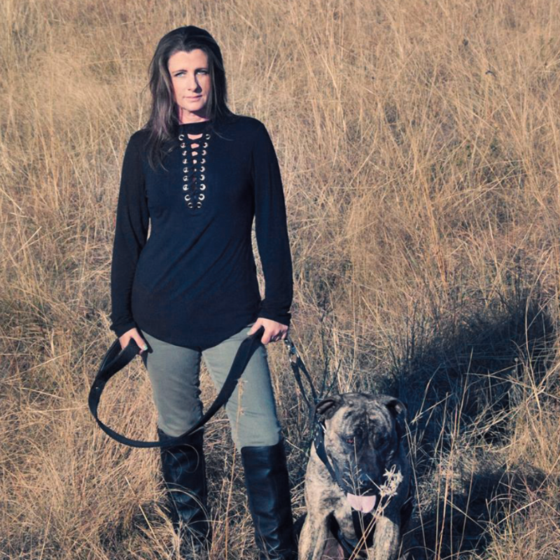 A woman standing in a field of tall, dry grass holding the leash of a brindle-colored dog.