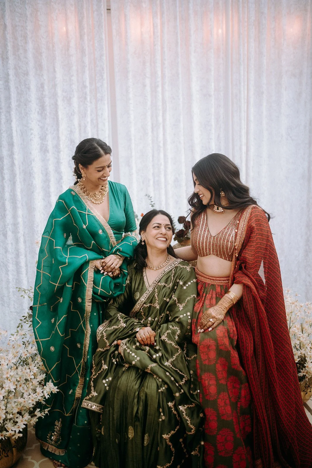 Three women in traditional Indian attire smiling and enjoying each other's company at an indoor event with white draped background and floral decorations.