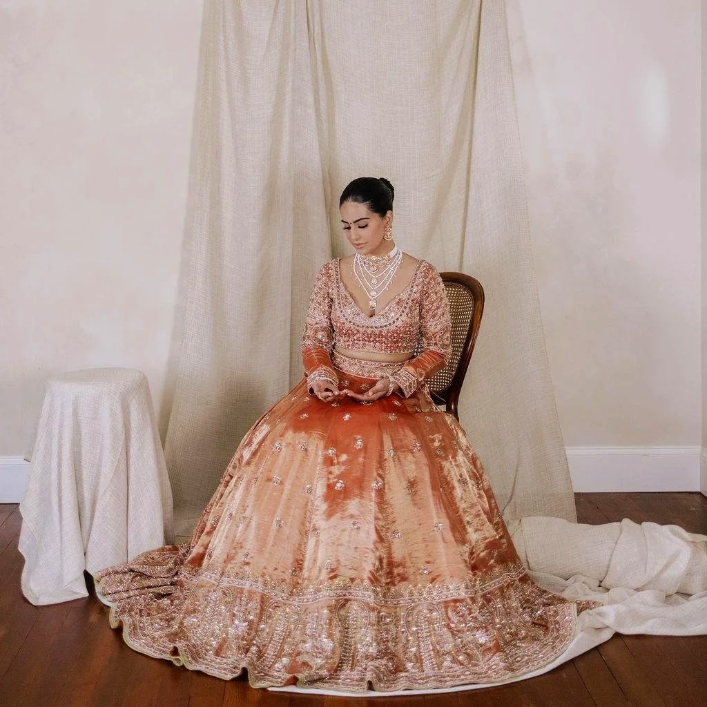 A woman in a traditional peach and gold Indian bridal lehenga sitting on a chair, looking down and holding her phone, with a beige curtain backdrop and a small table covered with cream cloth beside her.