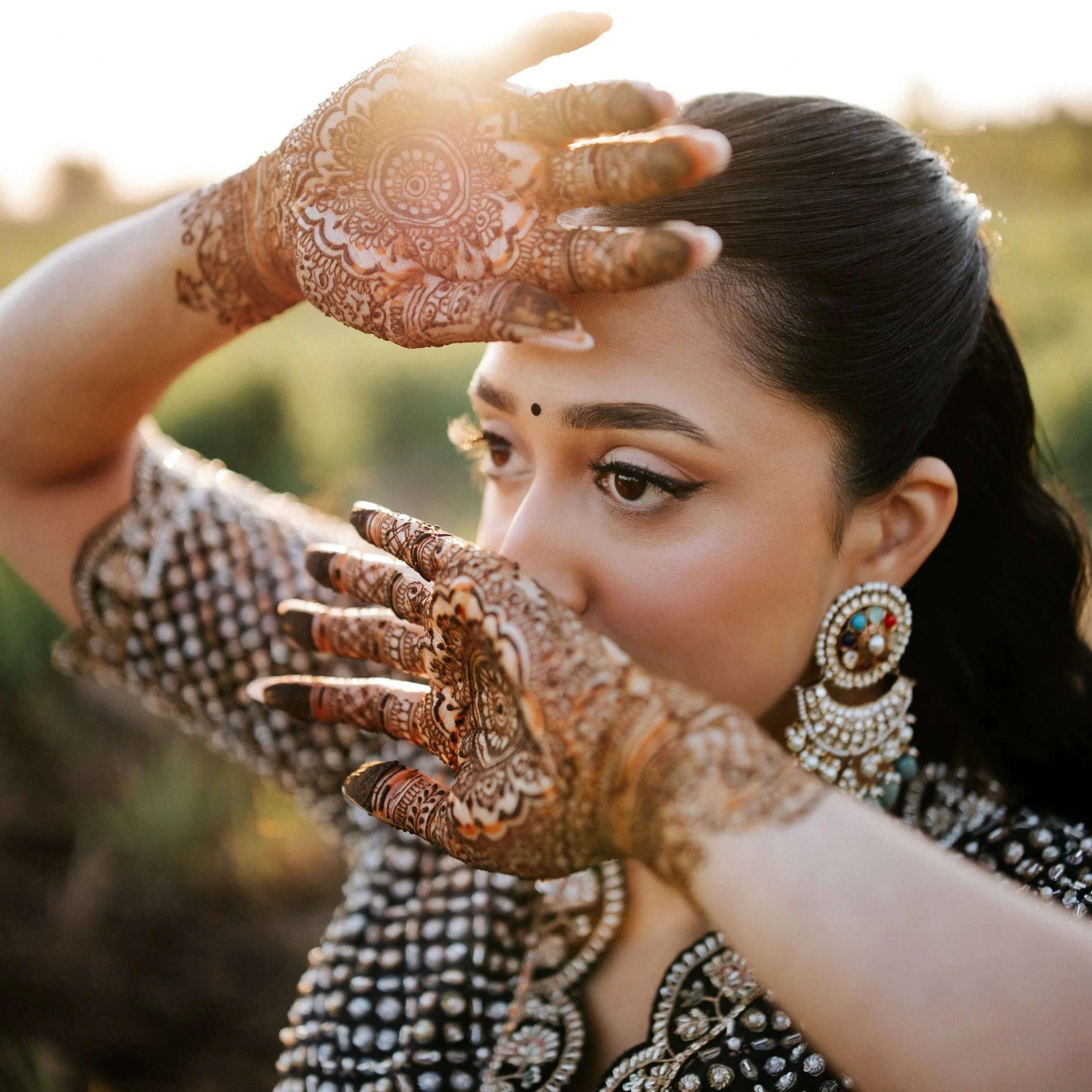 A woman with henna decorated hands, wearing traditional Indian jewelry, covering part of her face with her hand, outdoors at sunset.