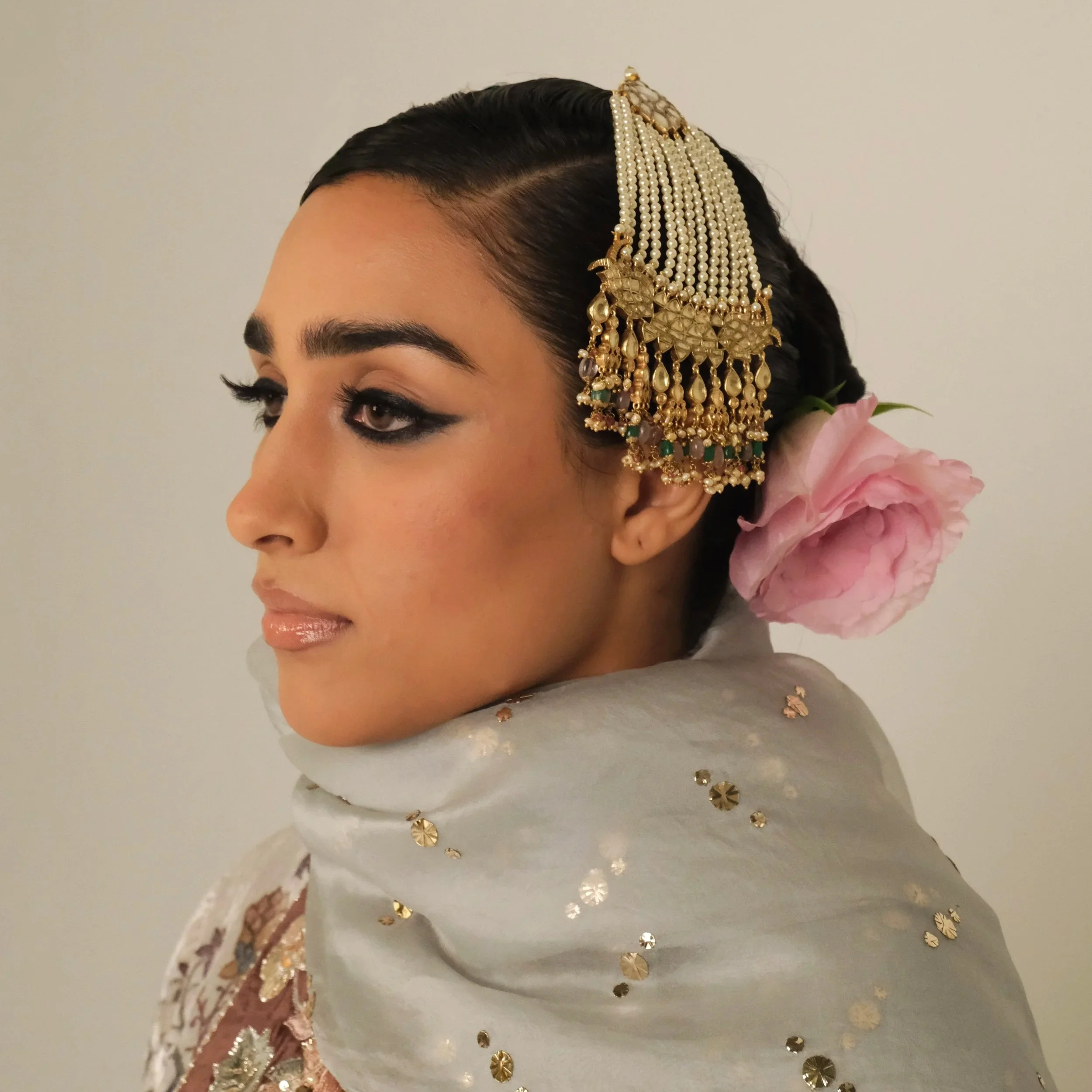 A woman with traditional Indian attire and jewelry, including a large beaded hair accessory, with a pink flower in her hair, wearing a white shawl adorned with gold accents.