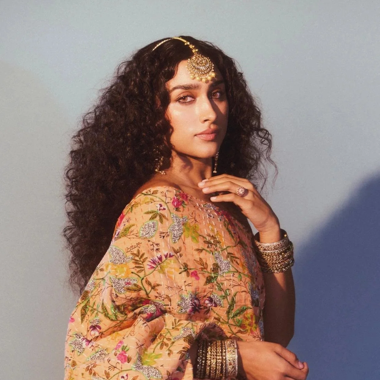 A woman with curly hair wearing traditional Indian jewelry, including earrings and a forehead ornament, and a floral saree, standing against a neutral background.