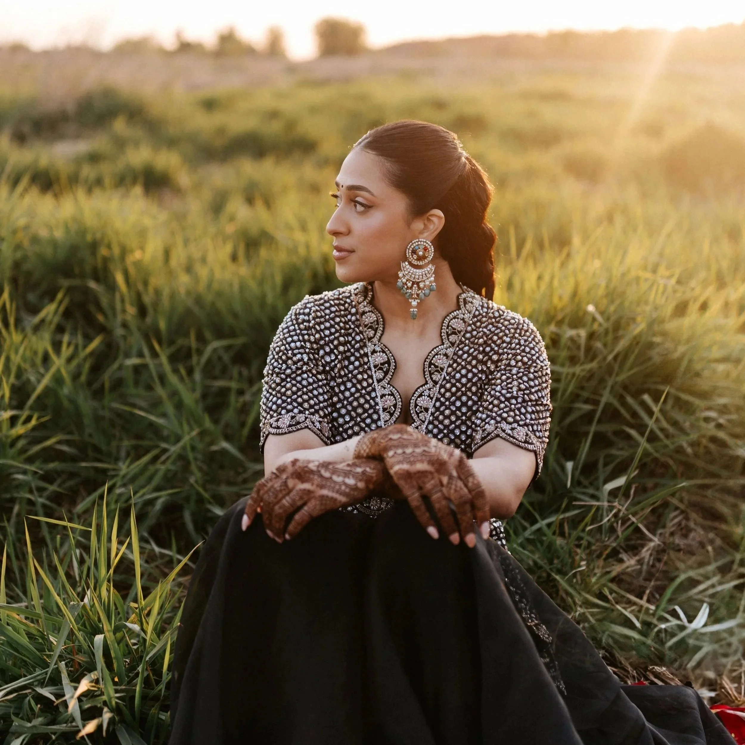 Woman dressed in traditional Indian attire sitting in a grassy field at sunset, with henna on her hands and wearing elaborate jewelry.