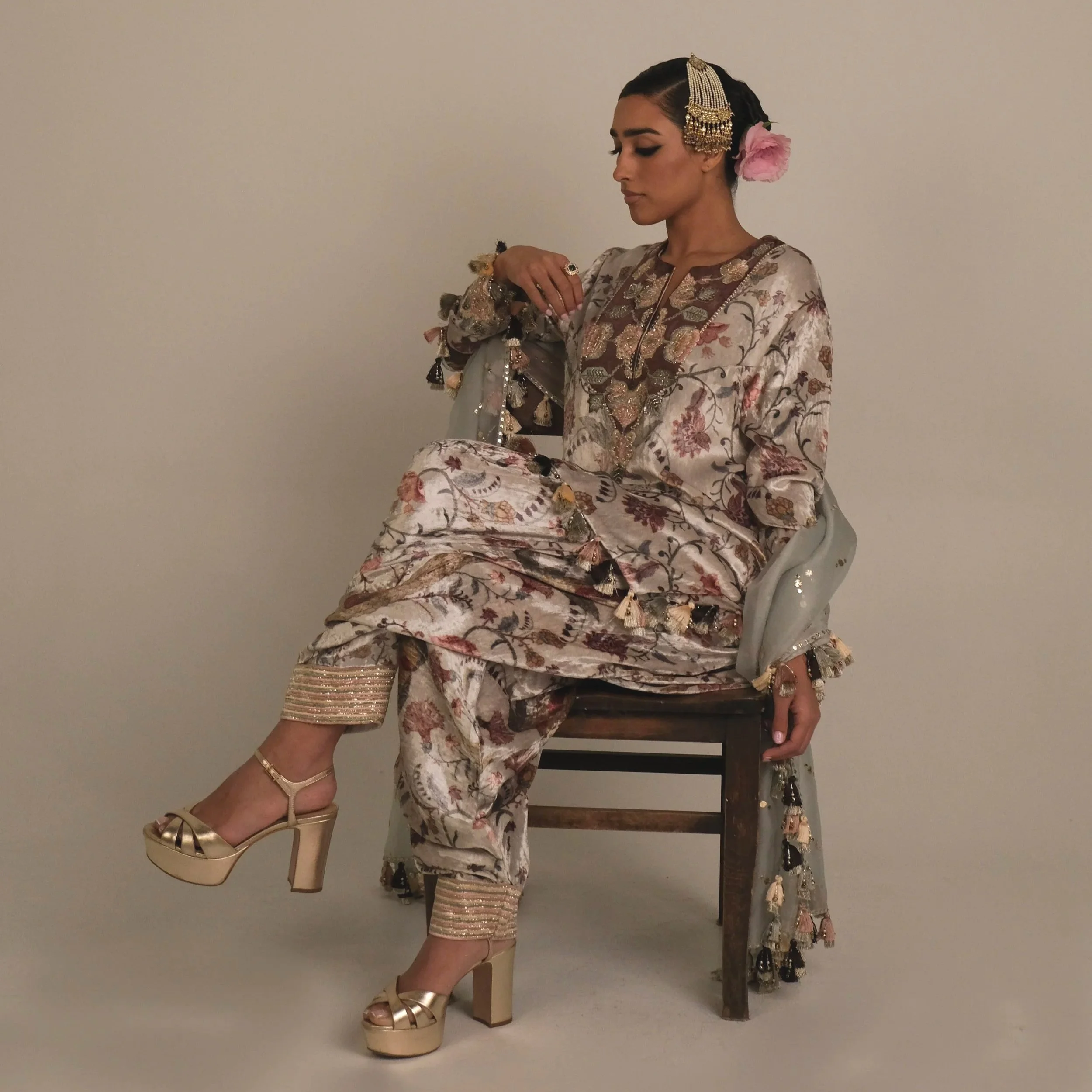 A woman dressed in traditional South Asian attire sitting on a wooden chair against a plain background, wearing a floral silk outfit with embellishments, high heels, and adorned with jewelry and a pink flower in her hair.