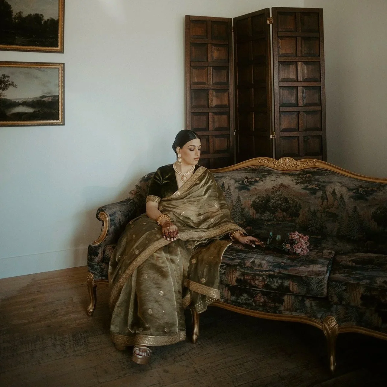 A woman dressed in traditional Indian attire sitting on a vintage floral sofa in a room with wooden flooring and a wooden room divider. She is wearing jewelry and has flowers in her hand.