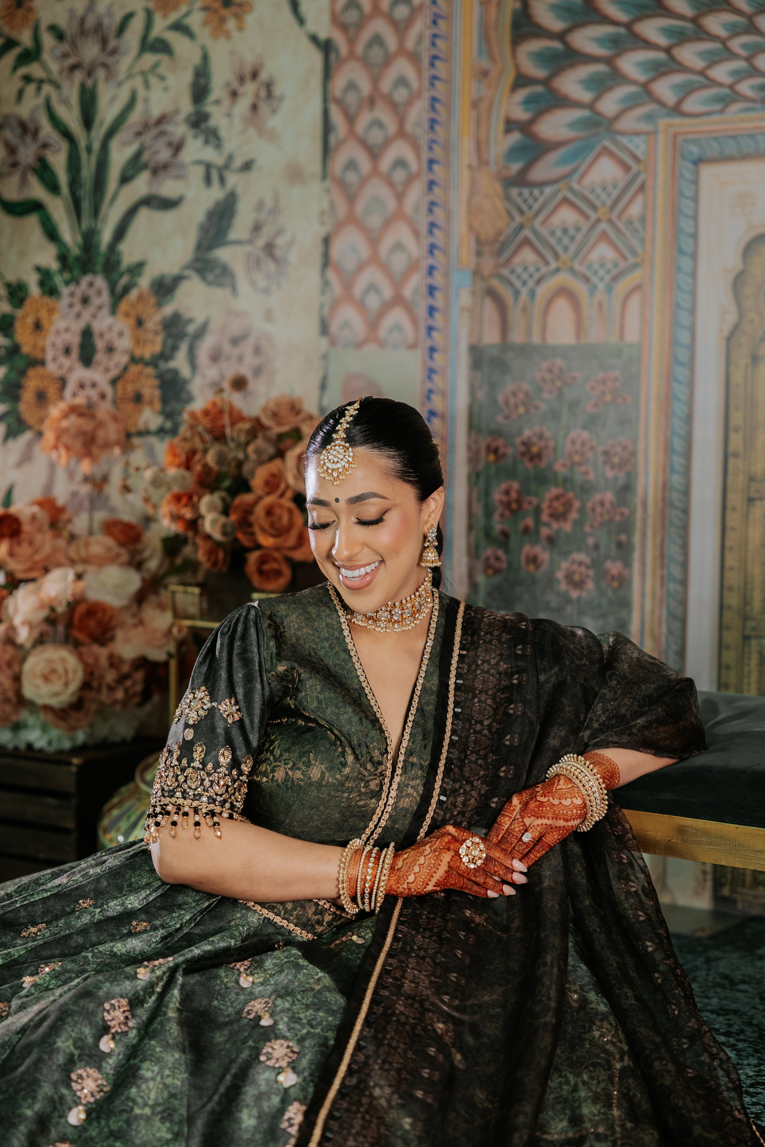 A woman dressed in traditional Indian attire sitting and smiling, with colorful floral background and ornate wall decorations.