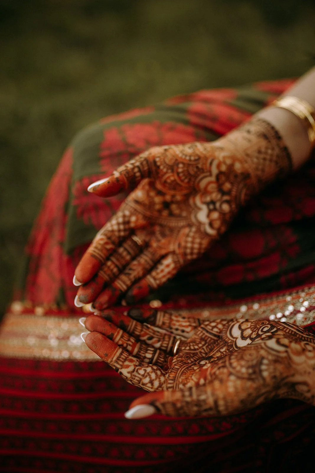 Close-up of a person's hand with intricate henna designs, resting on their lap, wearing traditional jewelry and a vibrant, patterned red and green outfit.