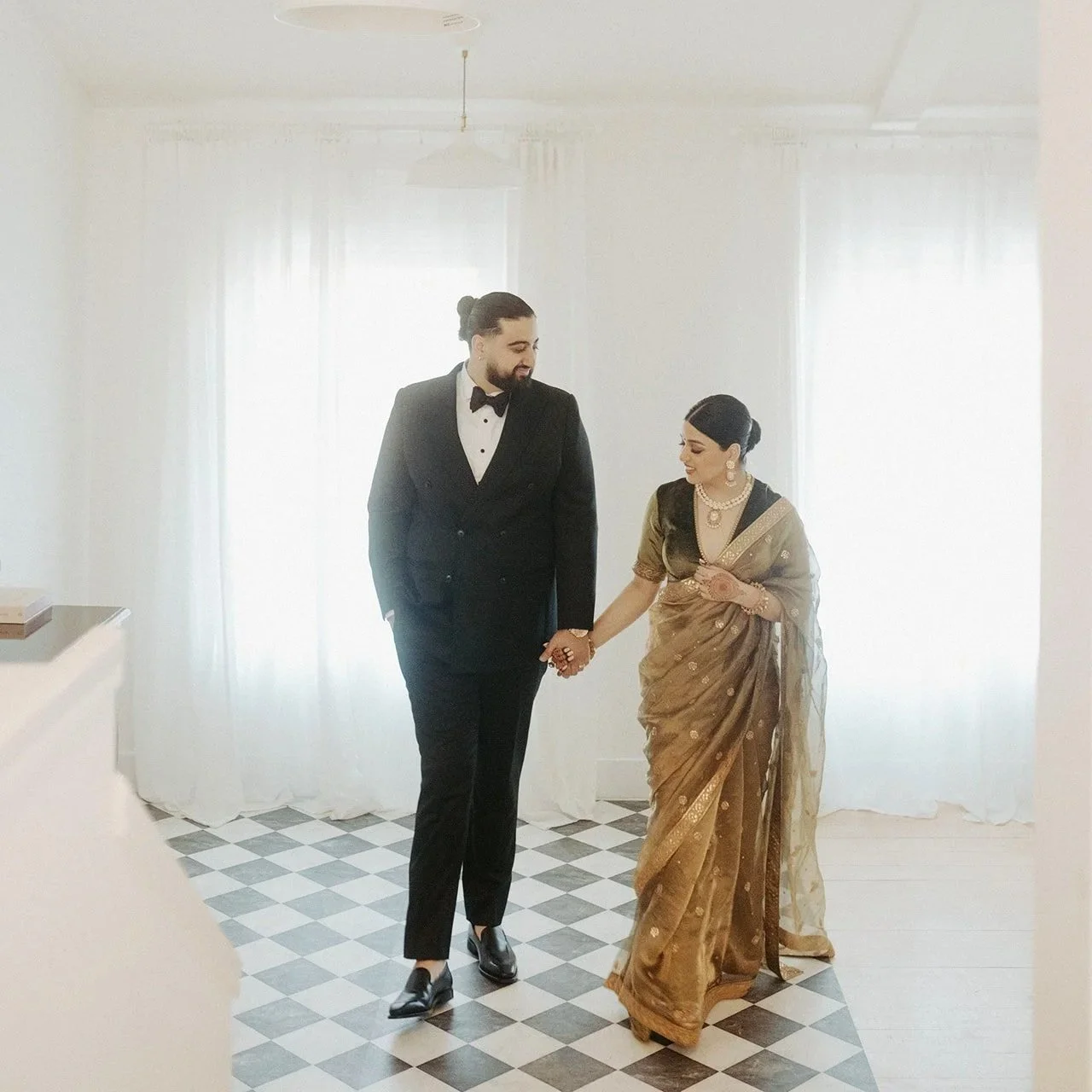 A couple dressed in formal attire walking hand in hand in a bright, white room with checkered black and white floors and sheer curtains.