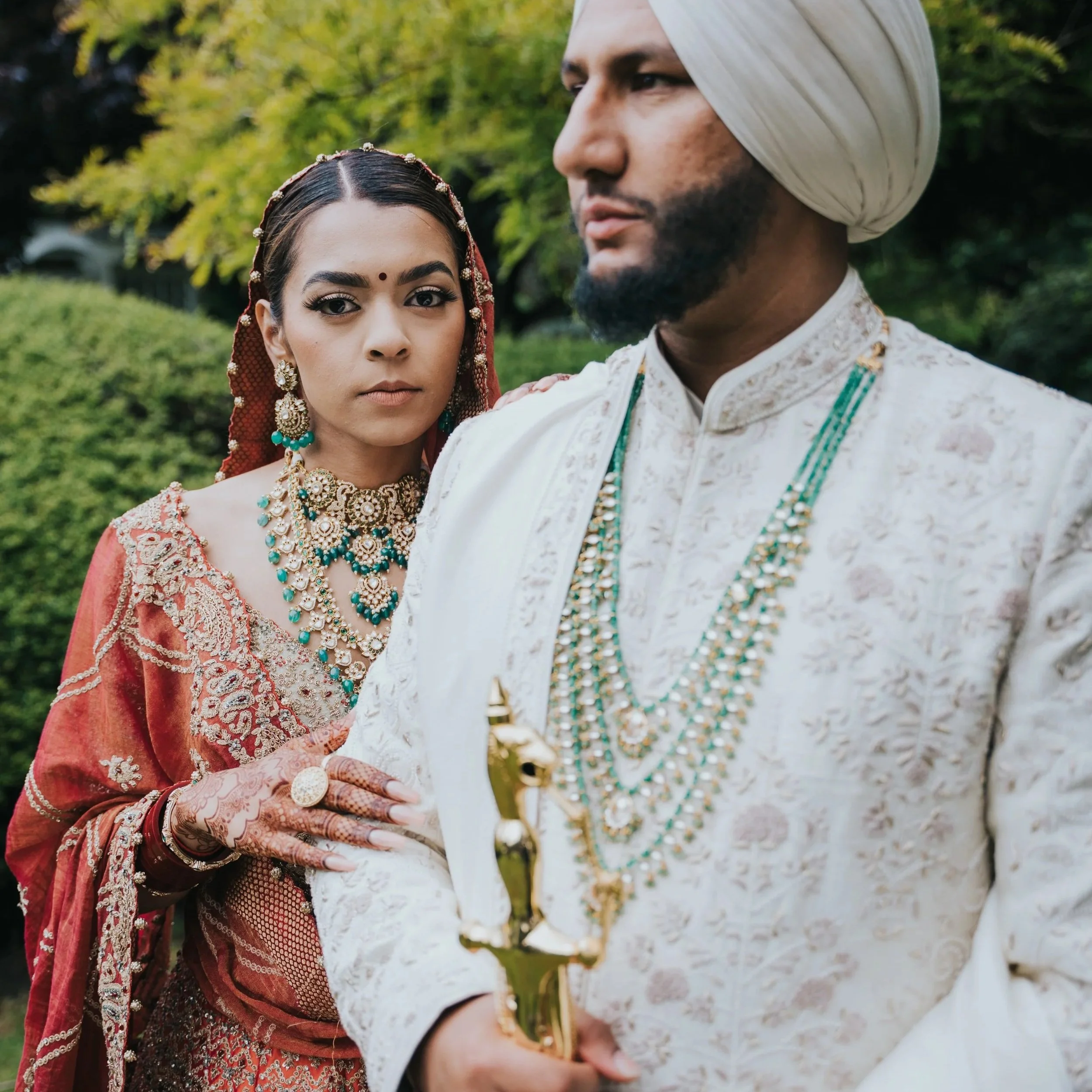 A couple in traditional Indian wedding attire, the woman adorned with jewelry and a red and gold outfit, and the man in an ivory sherwani with a turban, standing outdoors with greenery in the background.