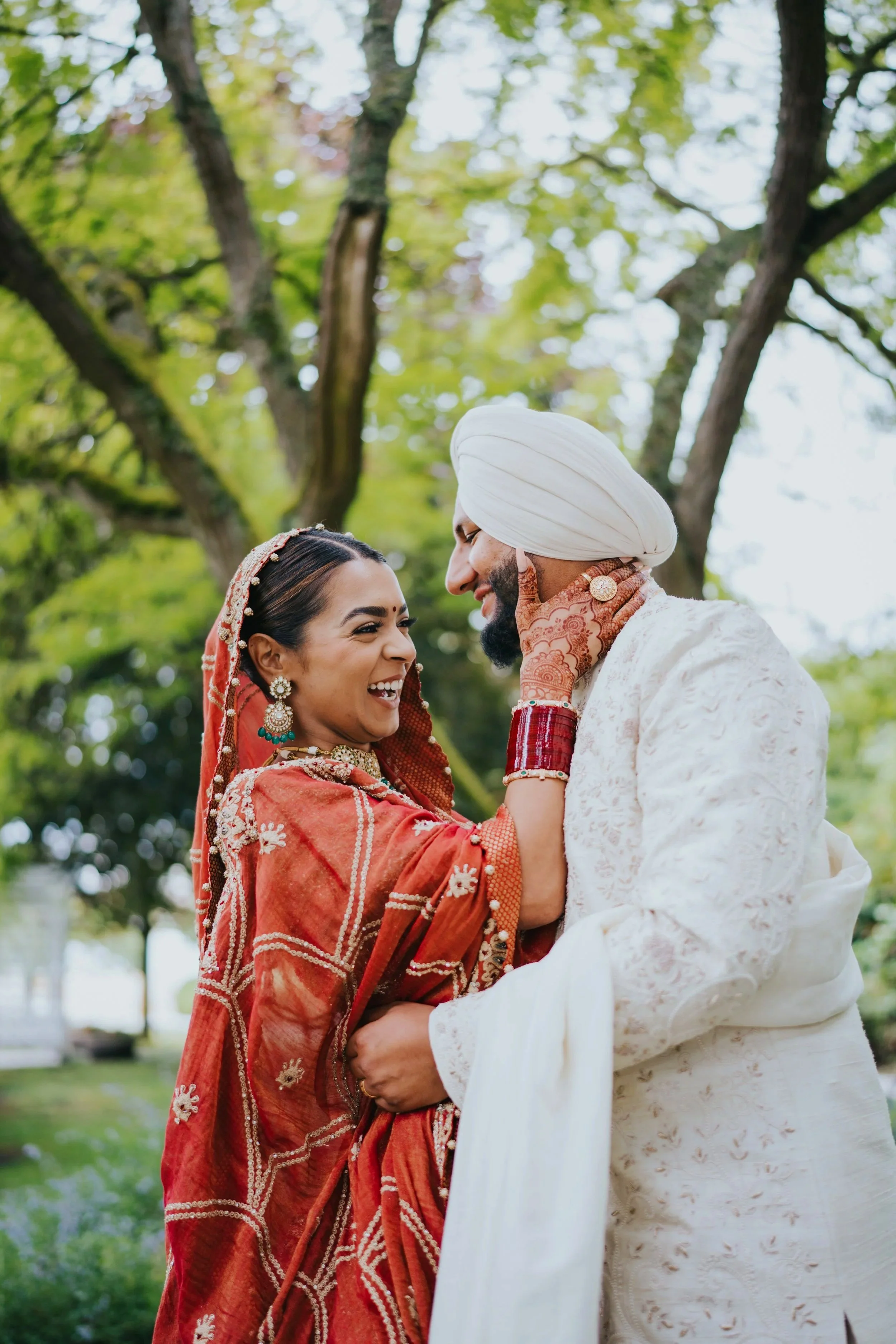A joyful Indian couple dressed in traditional wedding attire sharing a moment outdoors with trees in the background.