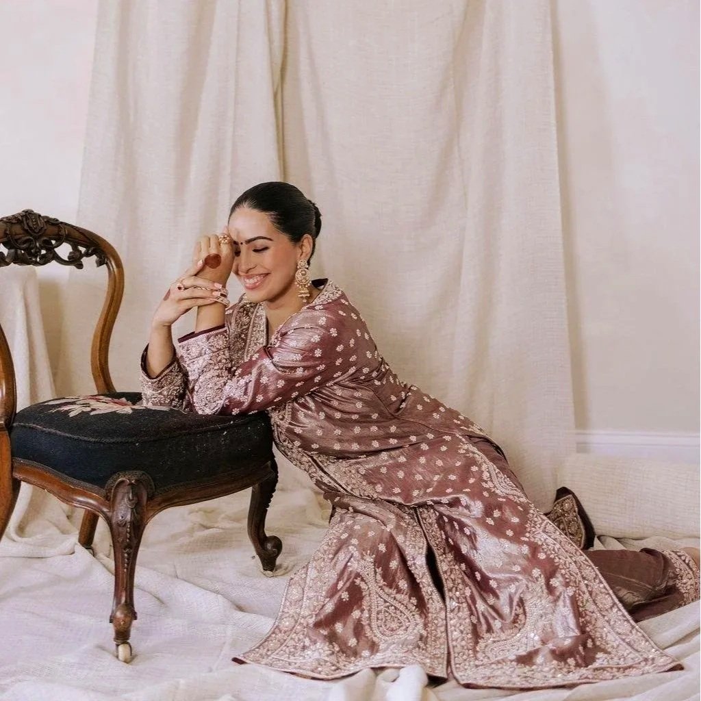 A woman in traditional attire sitting on the floor, leaning on a cushioned stool, smiling with closed eyes, with cream-colored curtains in the background.