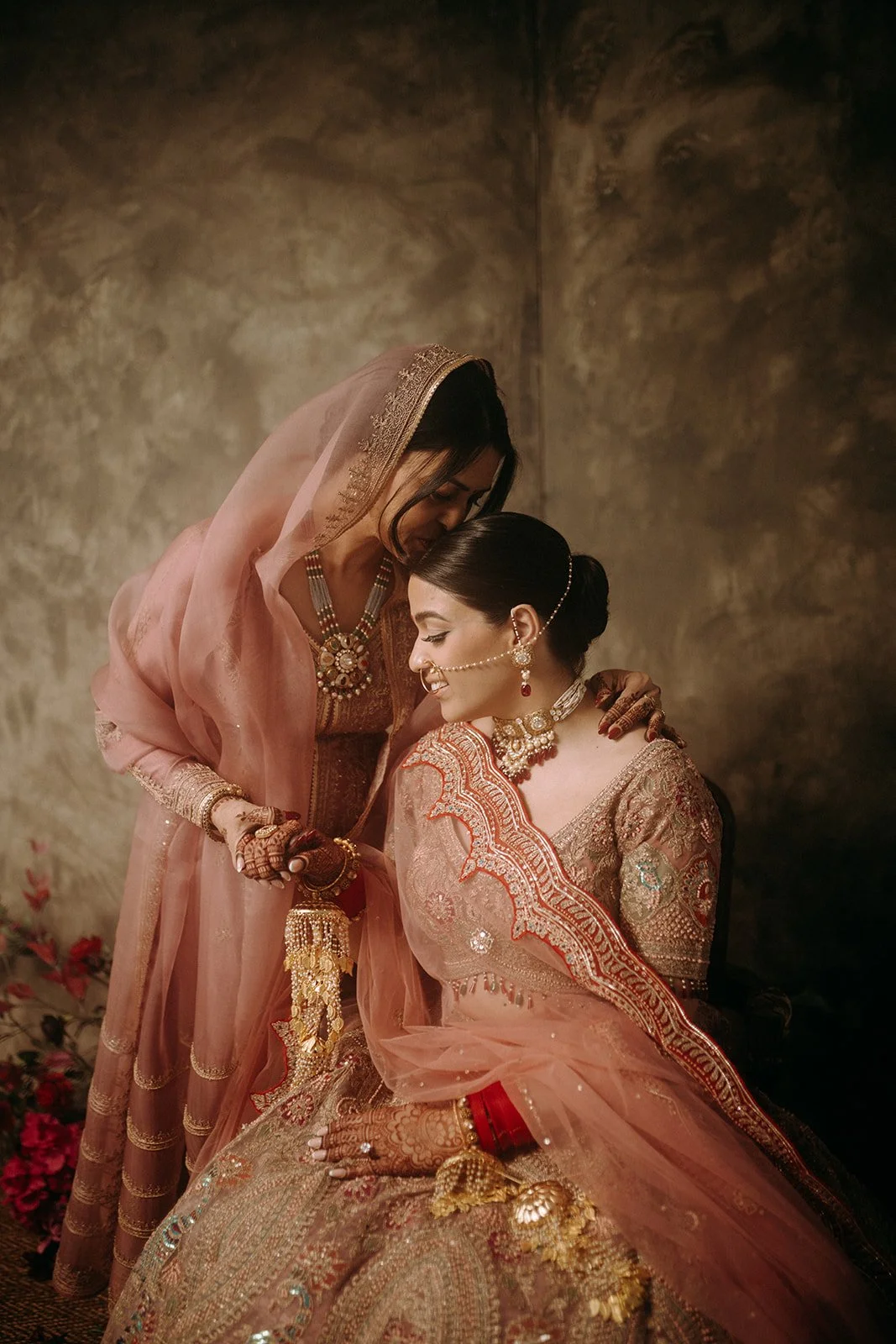Two women dressed in traditional Indian bridal attire, sharing a tender moment. One woman, seated, is wearing a heavily embellished sari with intricate embroidery, jewelry, and henna on her hands, while the other woman stands beside her, wearing an elegant sari with similar jewelry, holding her shoulder and leaning her forehead on her head in a loving gesture.