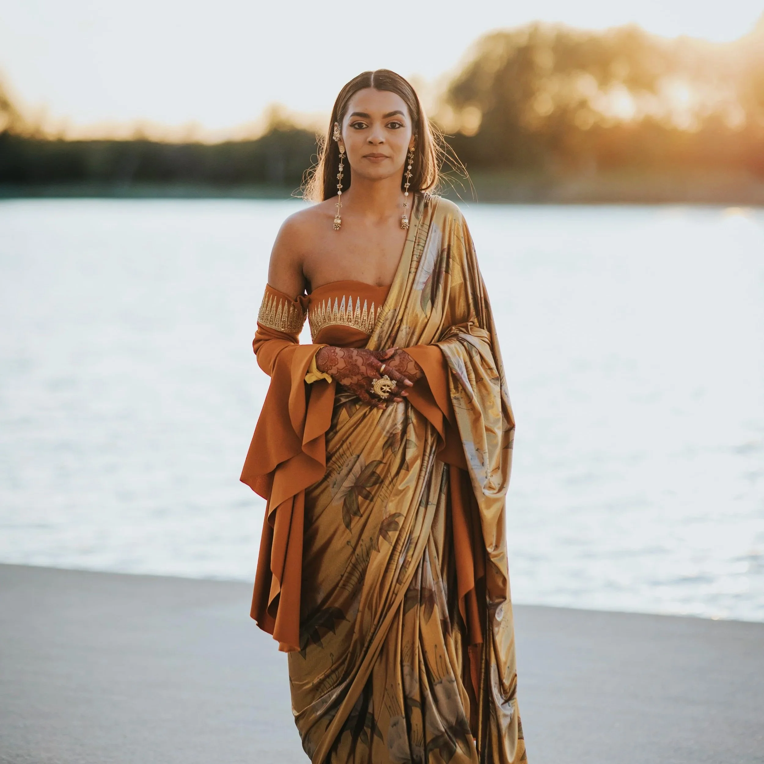 A woman wearing an elegant sari with intricate patterns and jewelry stands near a body of water during sunset.