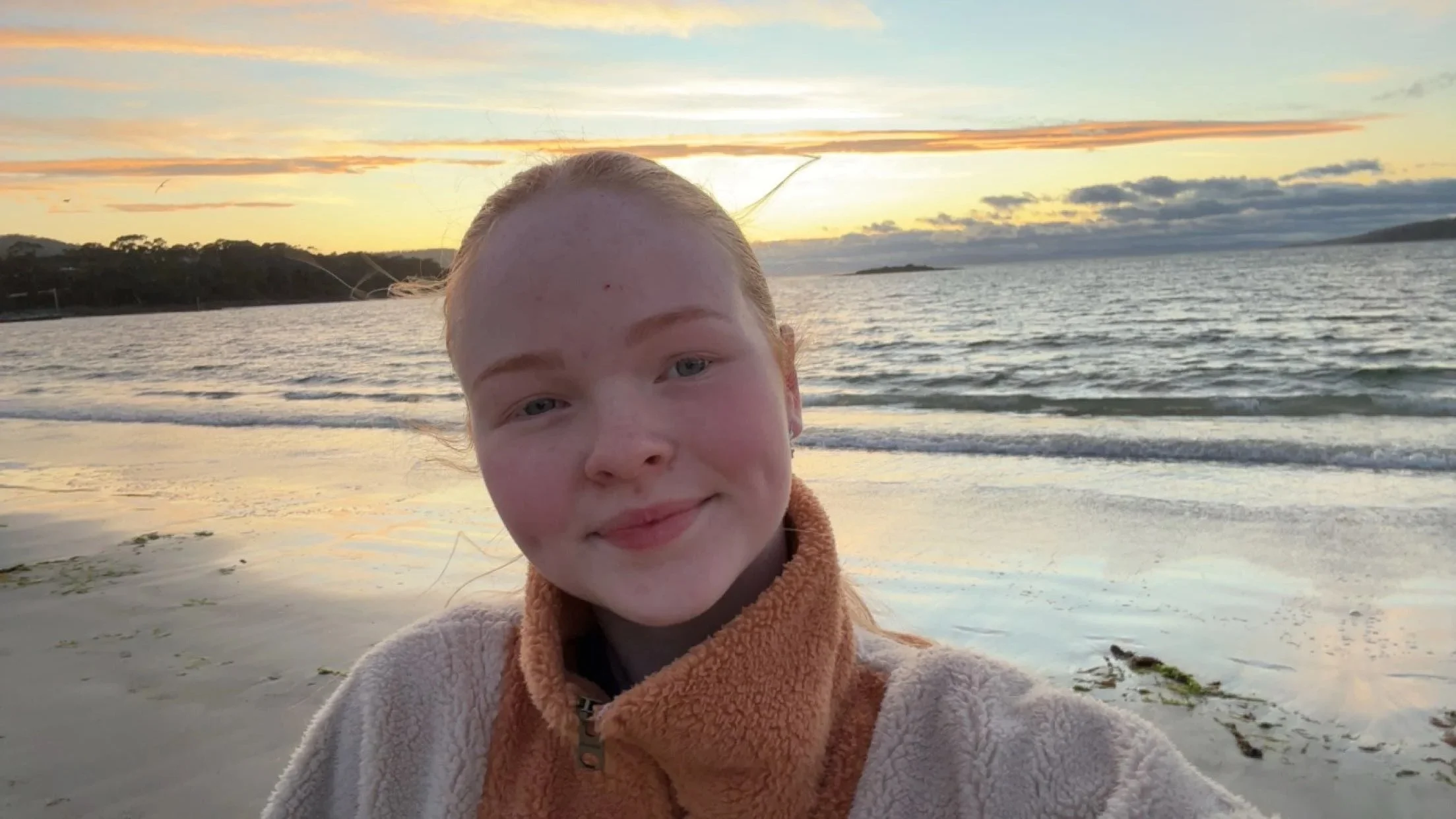 A young girl with red hair taking a selfie on the beach during sunset, with the ocean and sky in the background.