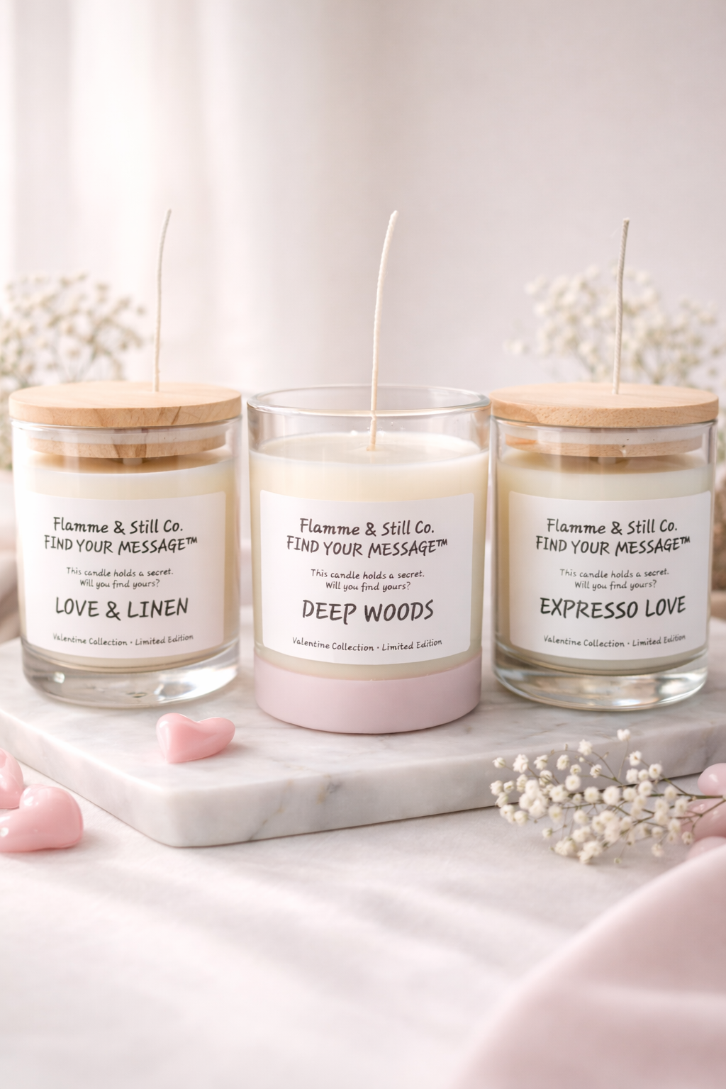 Three candles labeled 'LOVE & LINEN,' 'DEEP WOODS,' and 'EXPRESSO LOVE' from Flamme & Still Co. on a marble surface with pink heart-shaped objects and small white flowers around.