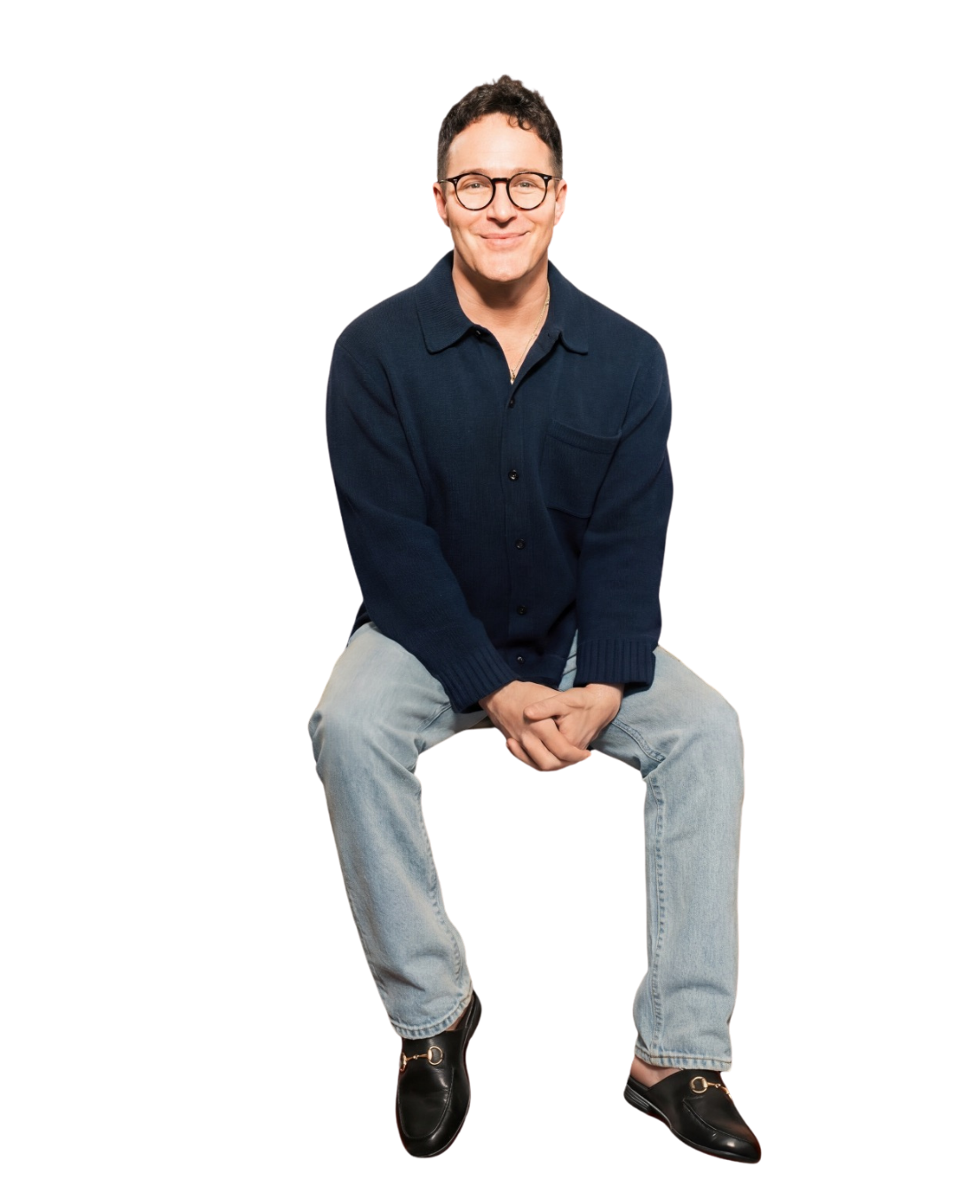 A man with glasses, wearing a navy sweater and light jeans, sitting with hands clasped, smiling at the camera.
