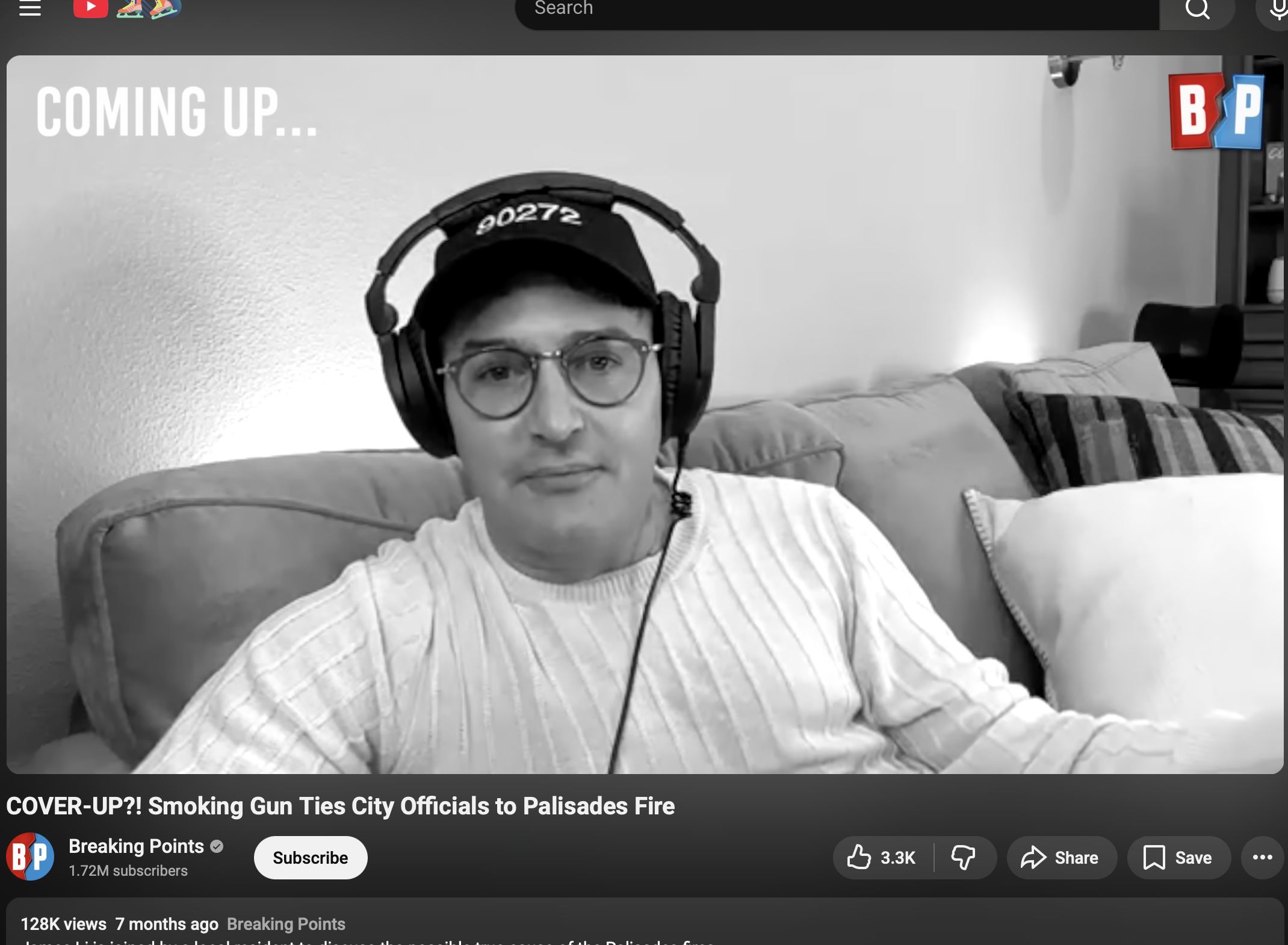 A man wearing glasses and a cap with headphones sits on a couch in a living room, appearing to record a video or speak to a camera. The background features a plain wall, a light source, and a shelf with decorative items. The video title indicates a news story about city officials and a fire.