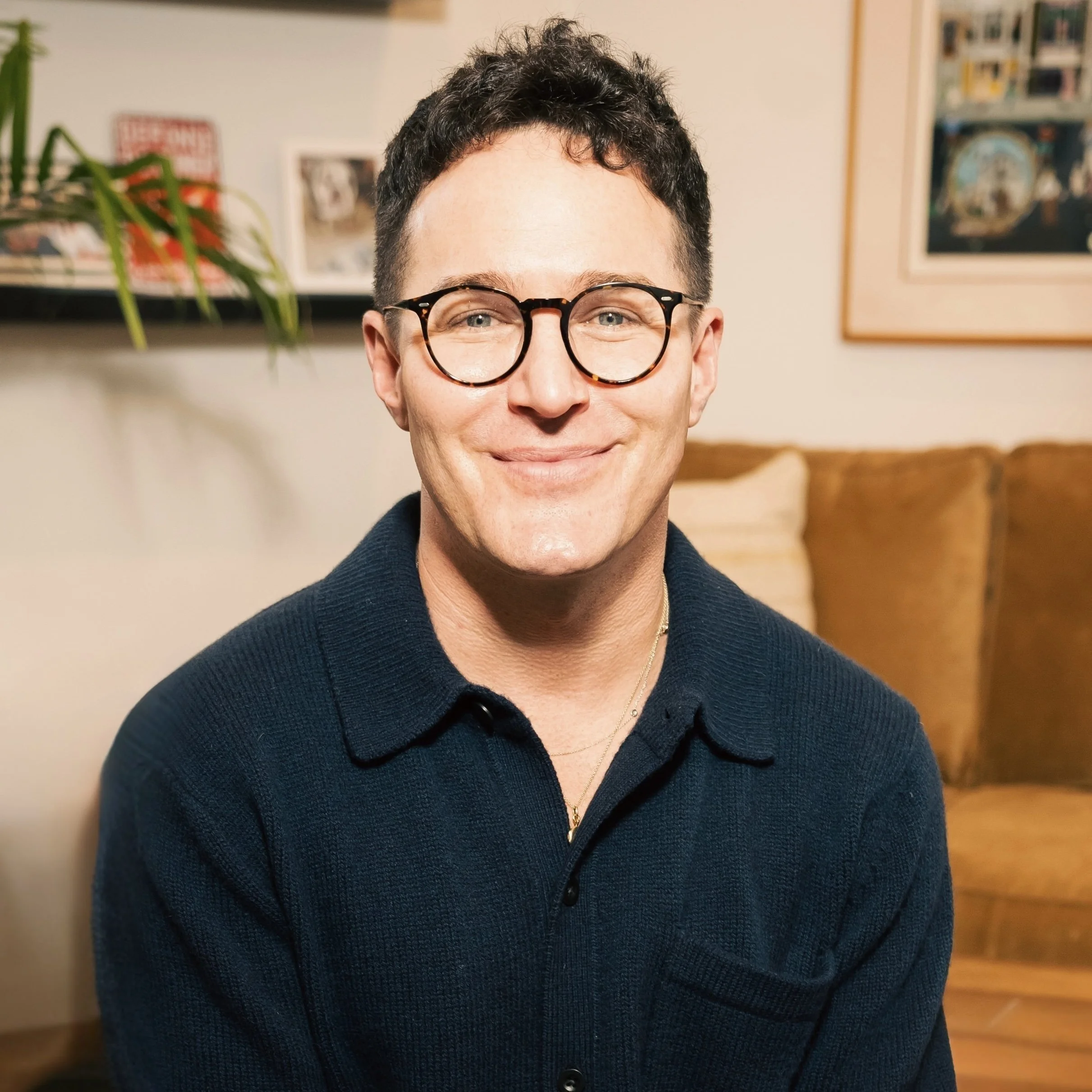 A smiling man with curly dark hair and round glasses sitting on a couch in a living room, with framed pictures hanging on the wall behind him.