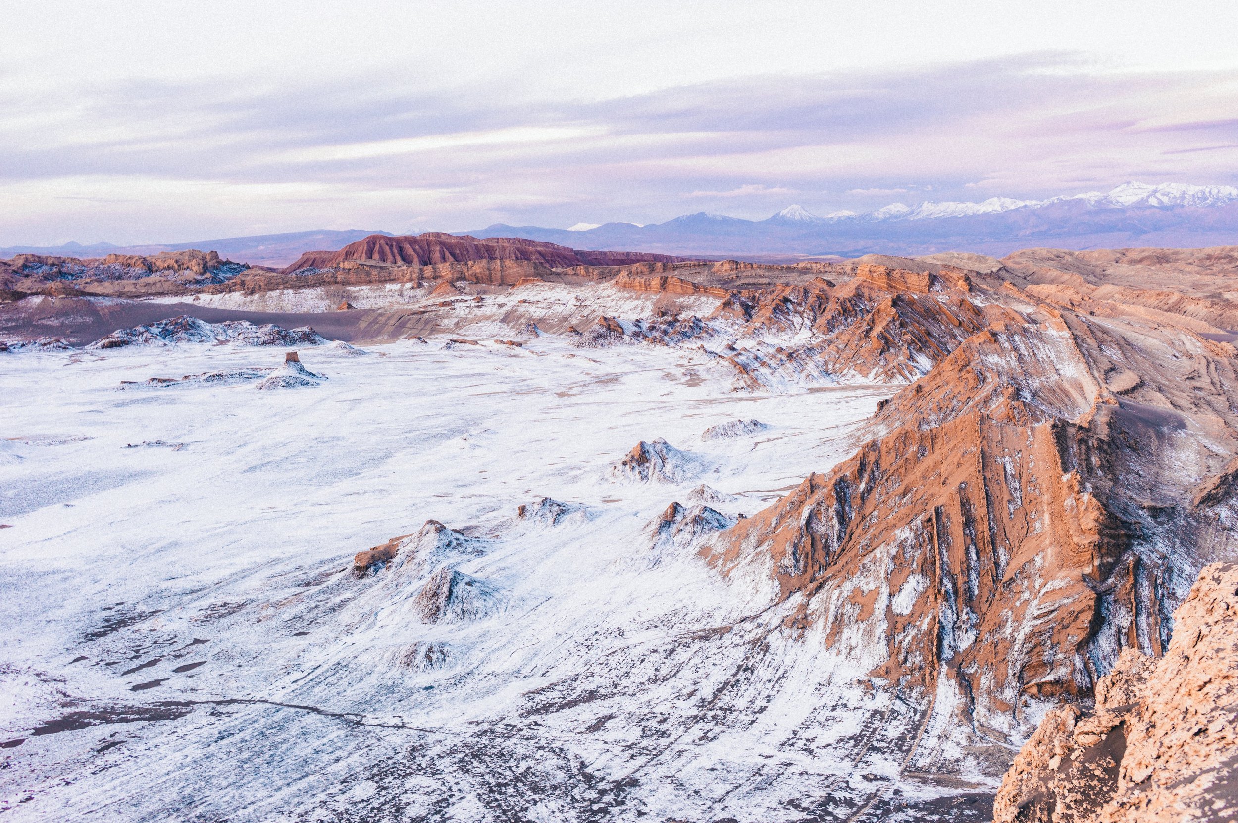 A mountainous desert landscape with snow-covered terrain and reddish-brown rocky formations, with distant snow-capped mountains under a cloudy sky.