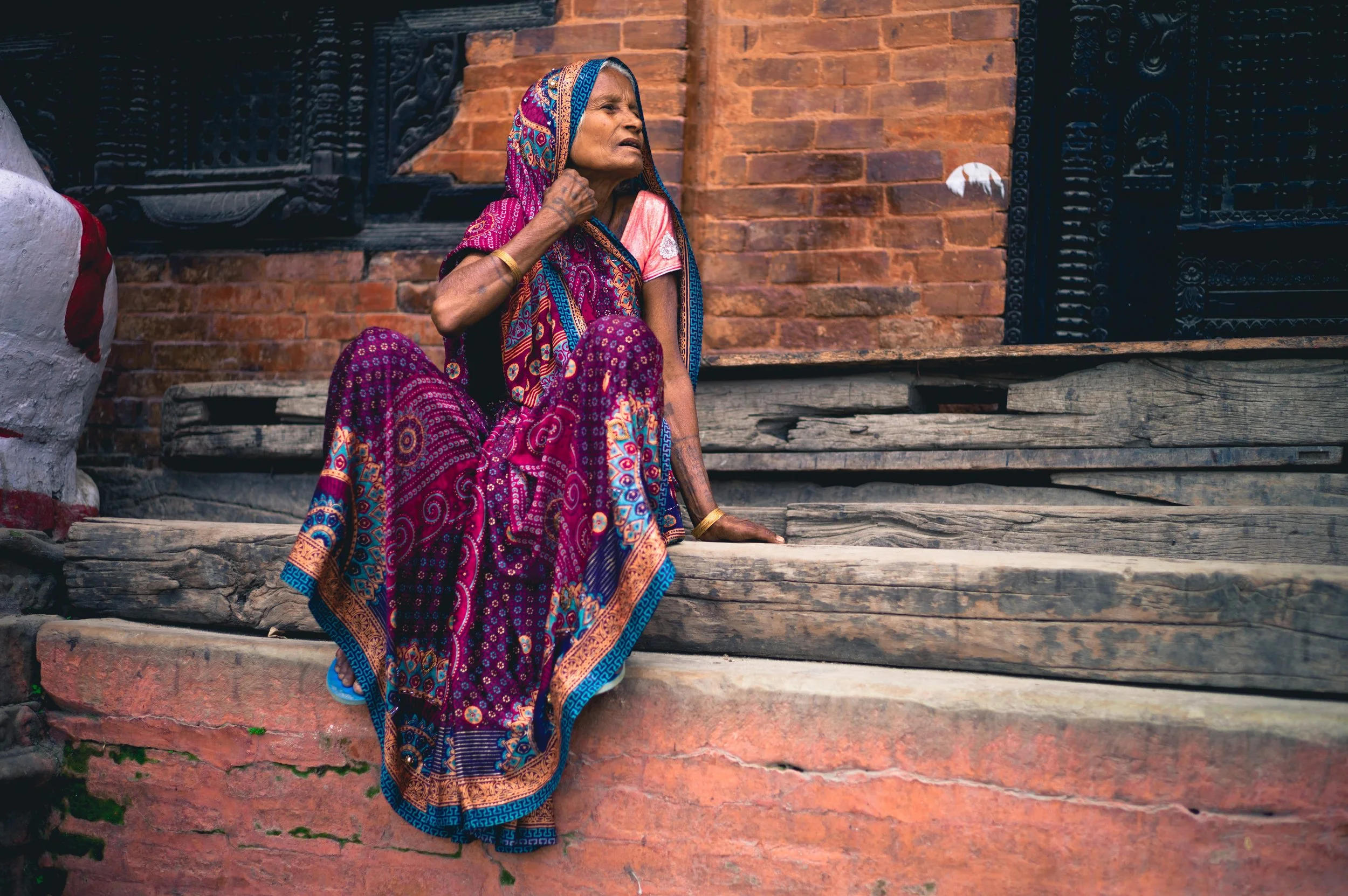 An elderly woman wearing a vibrant multicolored sari, sitting on wooden steps outside a brick building, looking to the side with a contemplative expression.