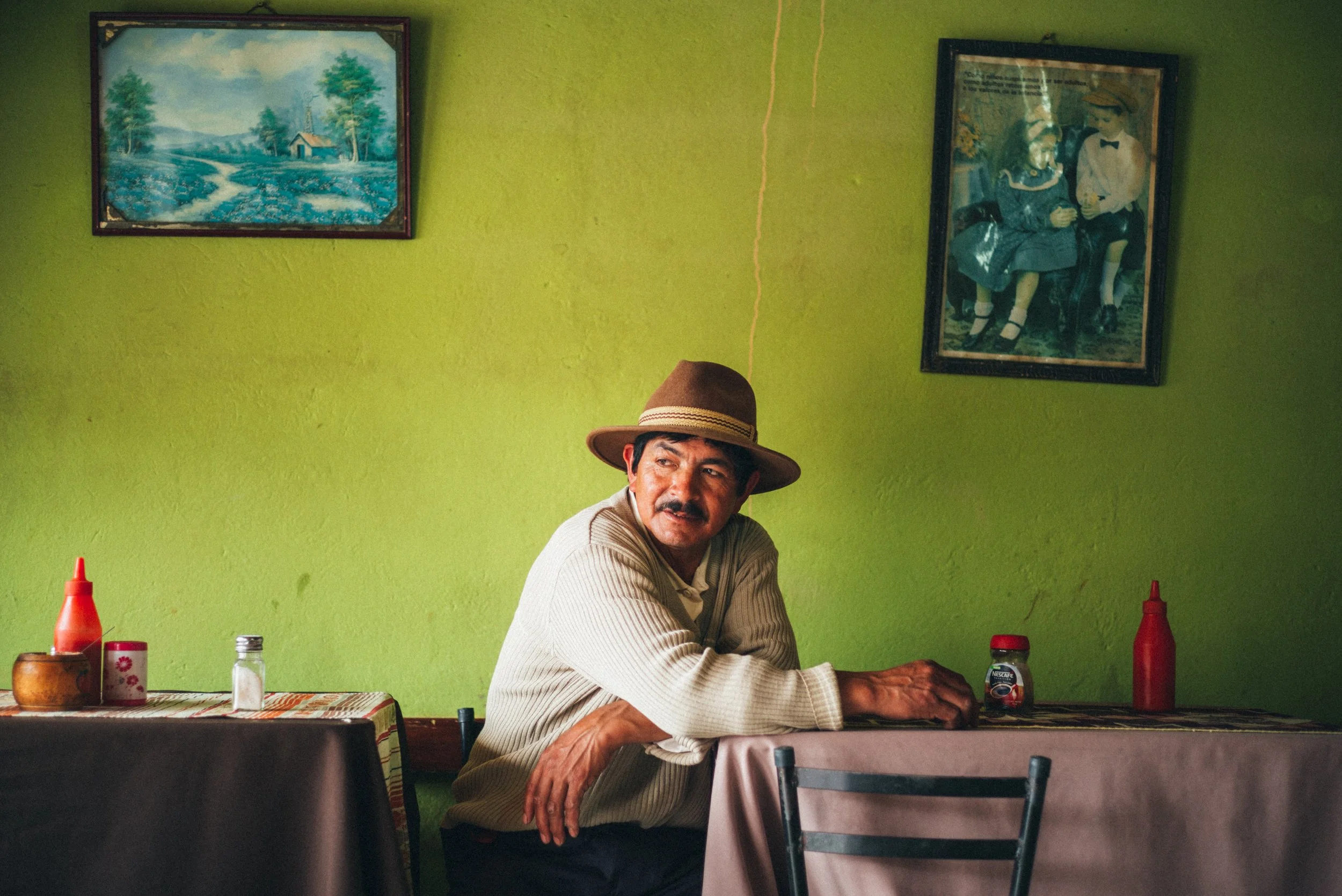 A man in a beige sweater and brown hat sitting at a table in a brightly lit room with green walls, decorated with two framed pictures, one showing a landscape and the other a painting of two children sitting together.