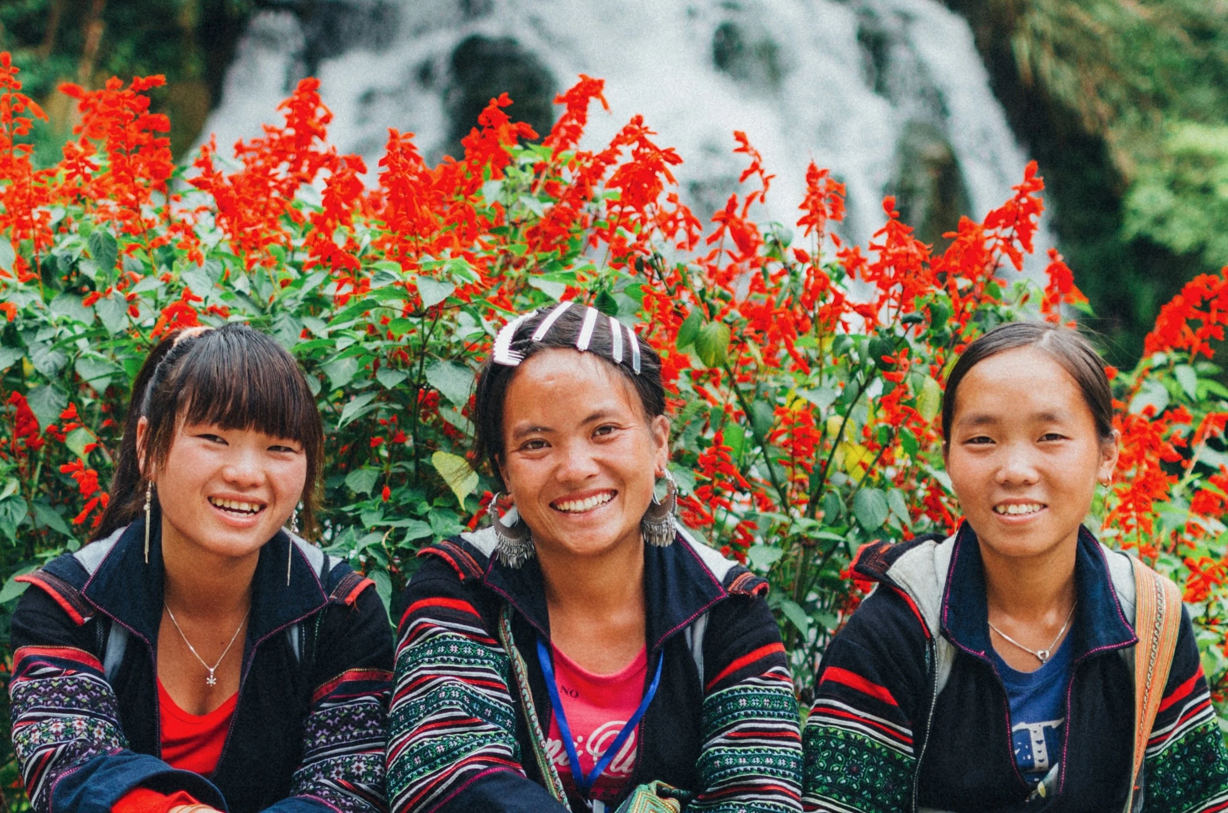 Three smiling women sitting outdoors in front of red flowers and greenery.